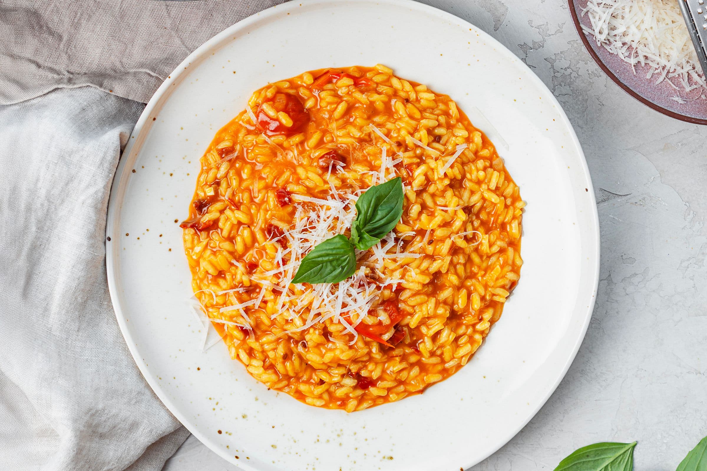 Vista de cima de um risotto cremoso de tomate num prato branco, coberto com parmesão vegan ralado e folhas de manjericão fresco.