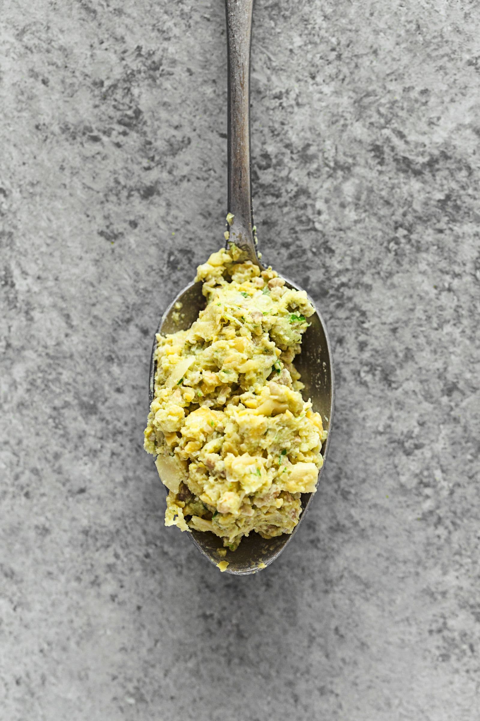 Uma colherada da pasta de grão e sementes de girassol sobre fundo cinzento.