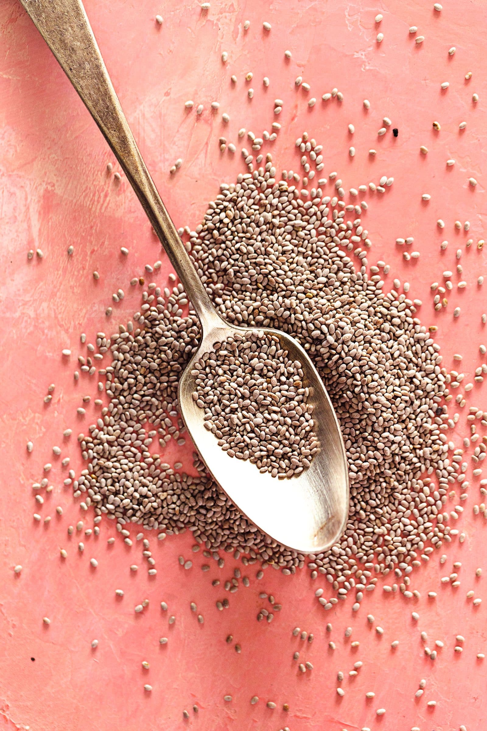 Colher cheia de sementes de chia brancas espalhadas sobre fundo rosa.