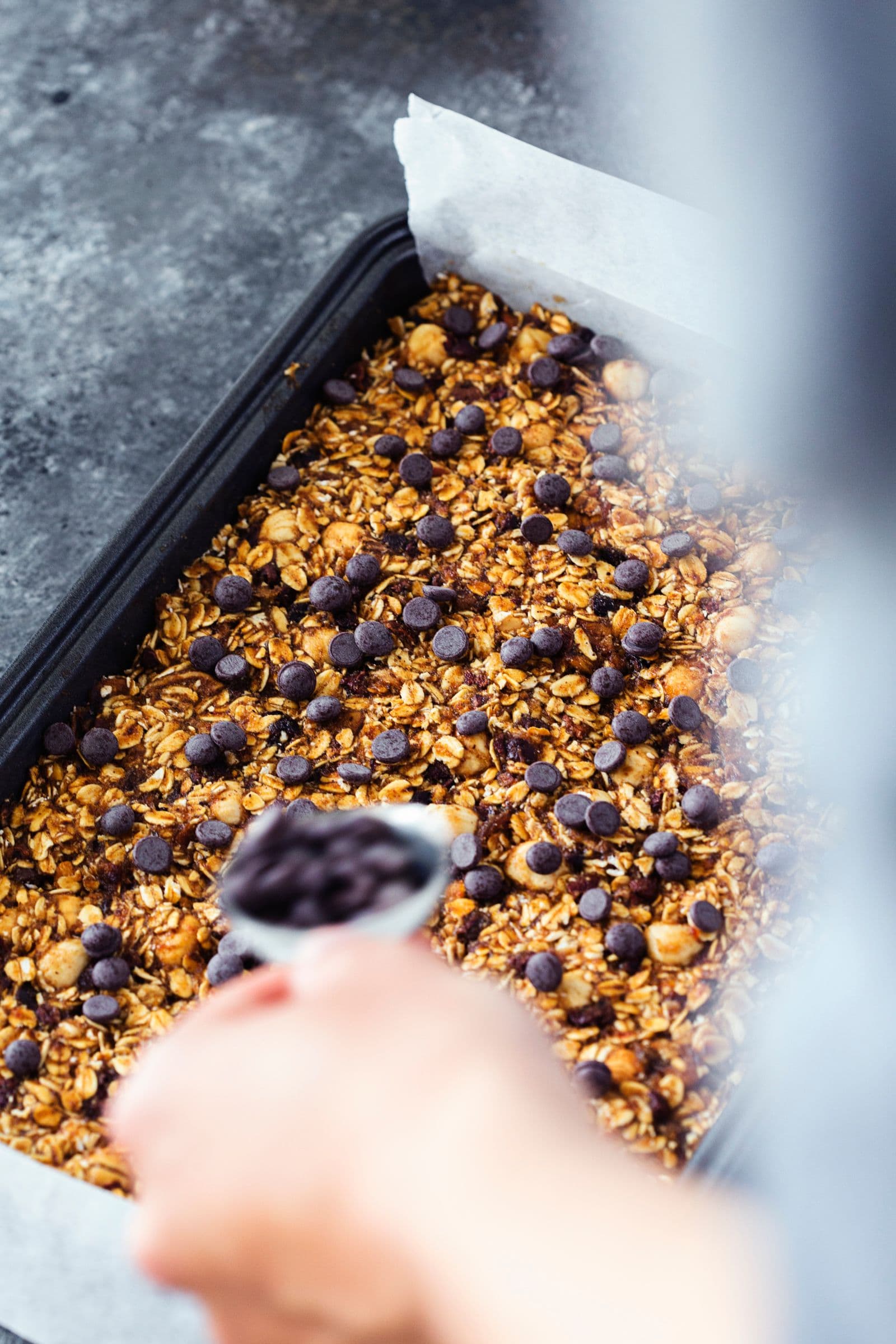 Mão a espalhar pepitas de chocolate negro sobre mistura de granola.