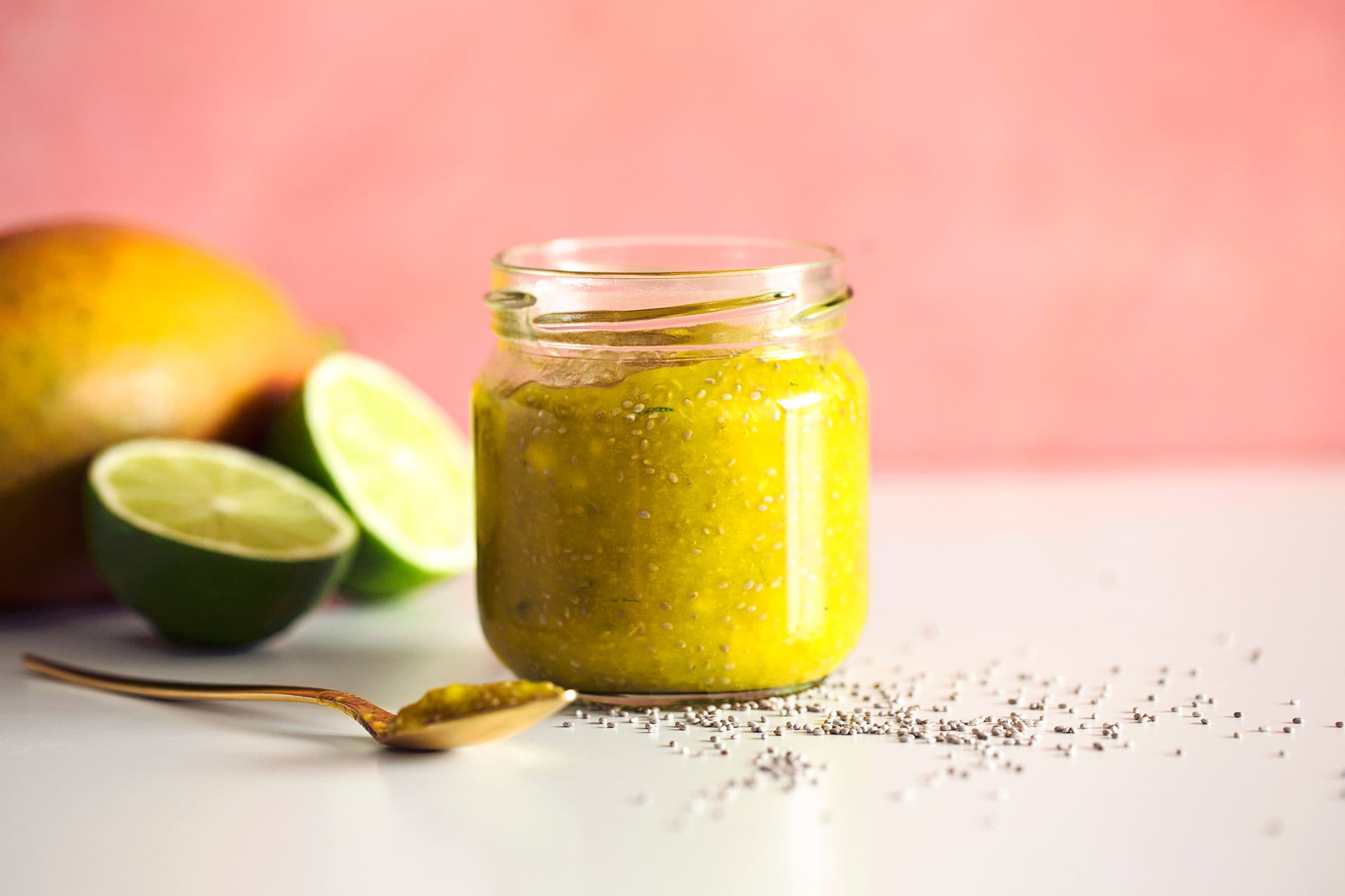 Jar of homemade quick mango chia jam and with lime halves and whole mango on a pink background.