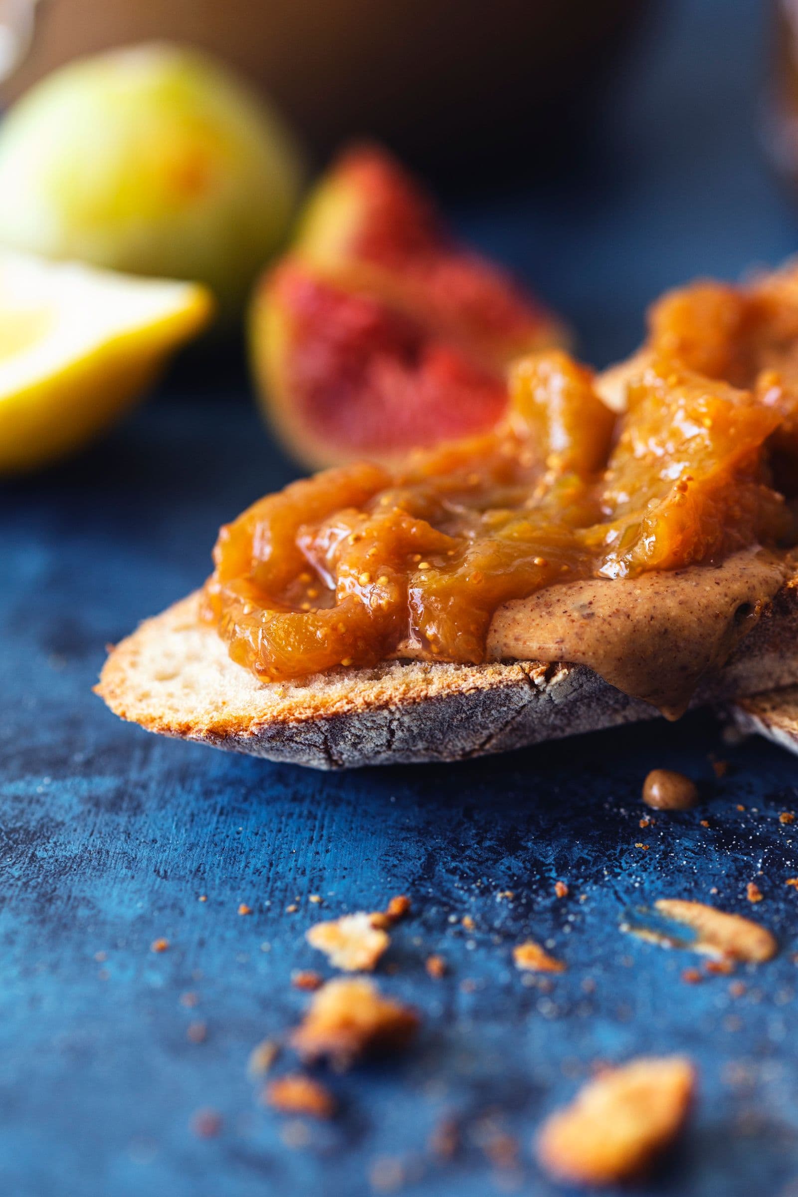 Tosta com manteiga de amêndoa e doce de figo, com migalhas e fruta ao fundo.