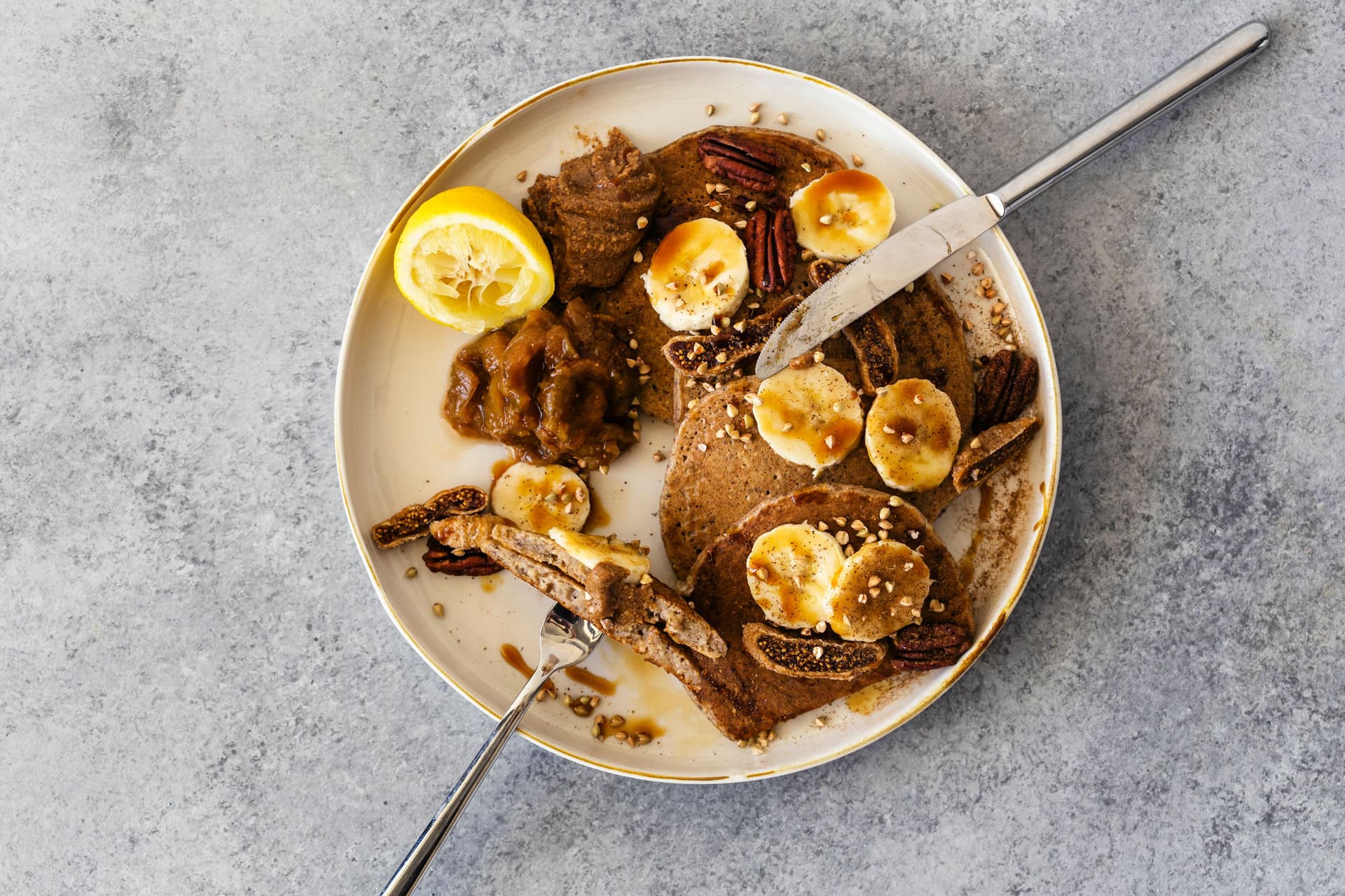 Prato de panquecas de aveia com banana e limão, cobertas com manteiga de amêndoa com especiarias chai, compota de figo e gengibre, rodelas de banana e nozes pecã.