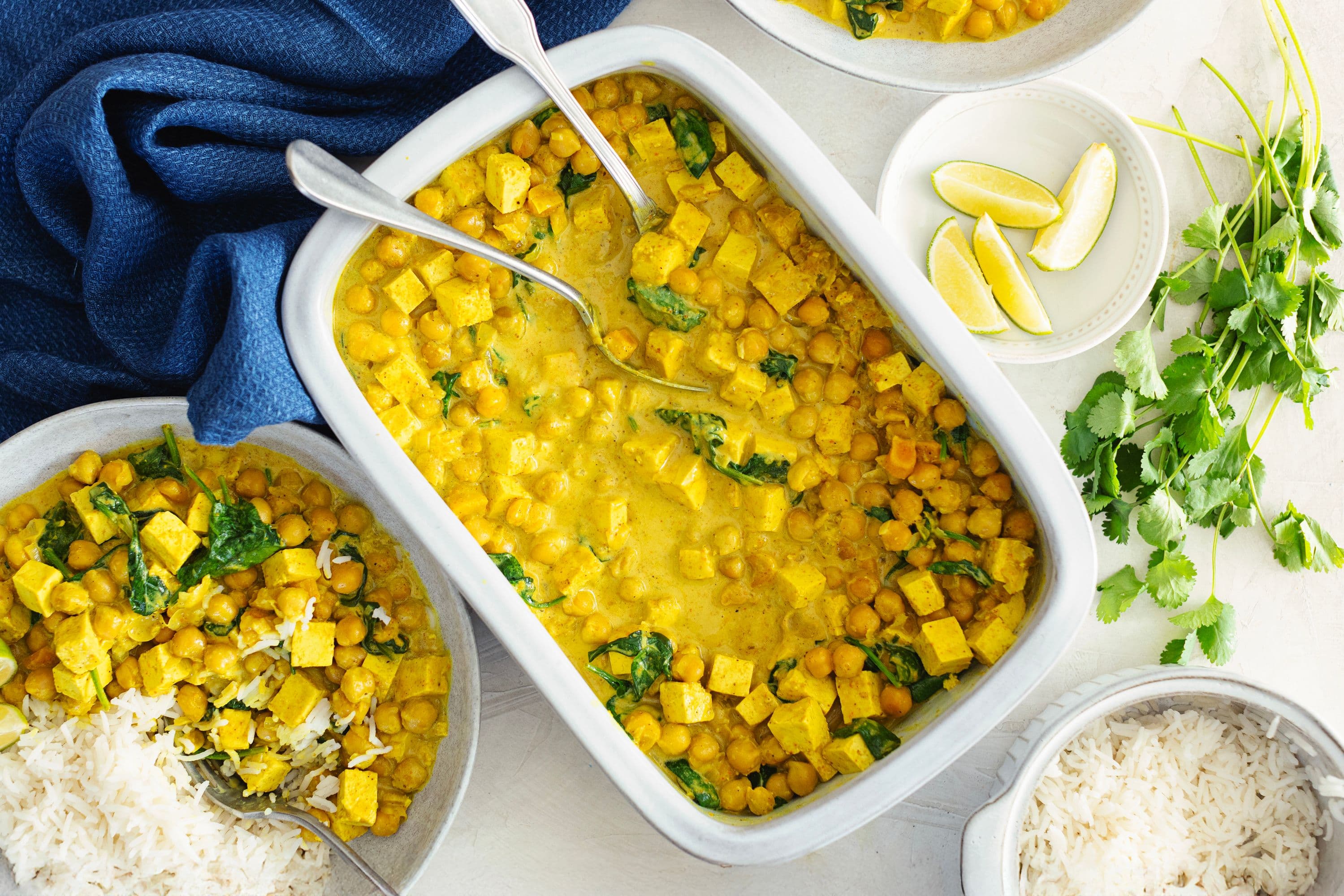White baking dish filled with chickpea, tofu and spinach curry alongside bowls of rice and lemon wedges.