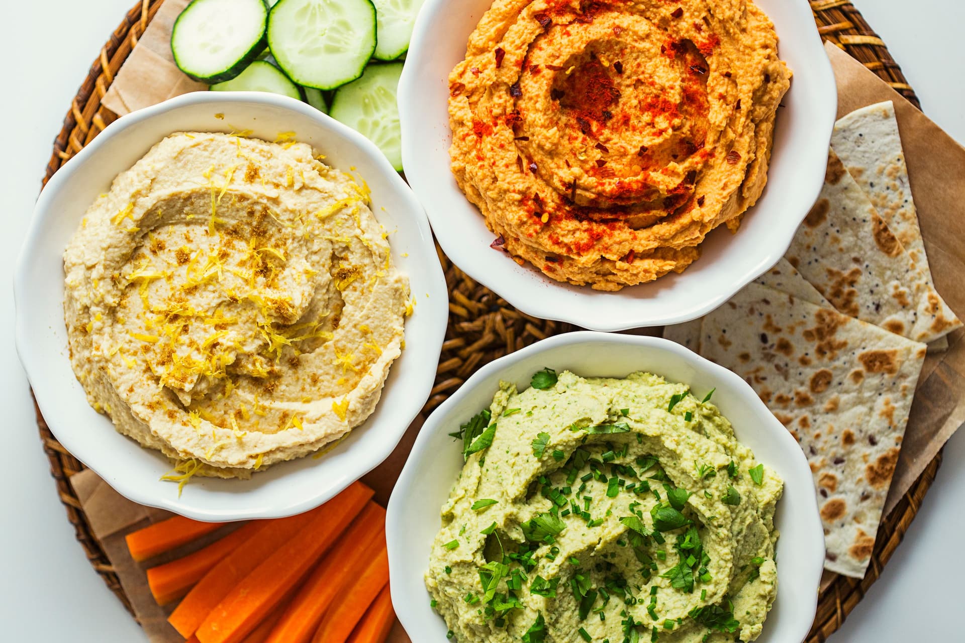 Três taças coloridas de húmus—limão, tomate e ervas—servidas com tortilhas e palitos de vegetais.