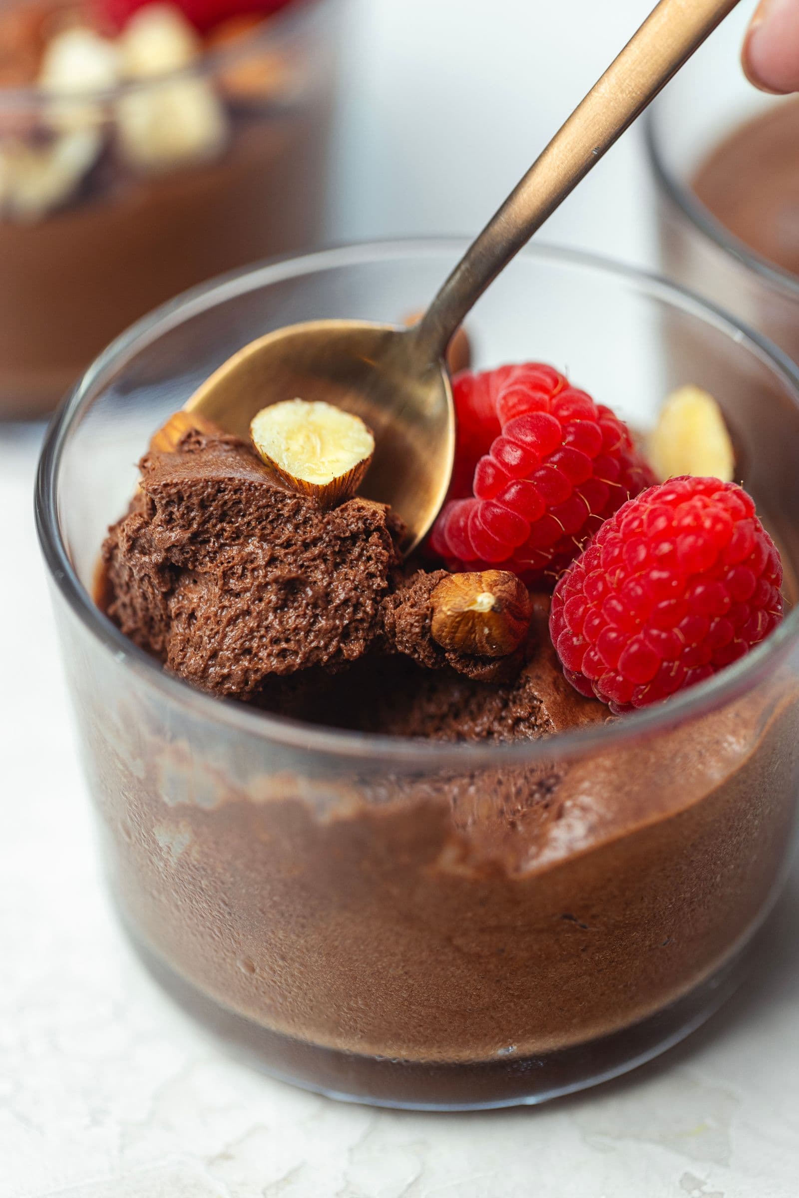 Colher a retirar uma porção de mousse de chocolate vegan fofa, num copo de vidro com framboesas e avelãs.