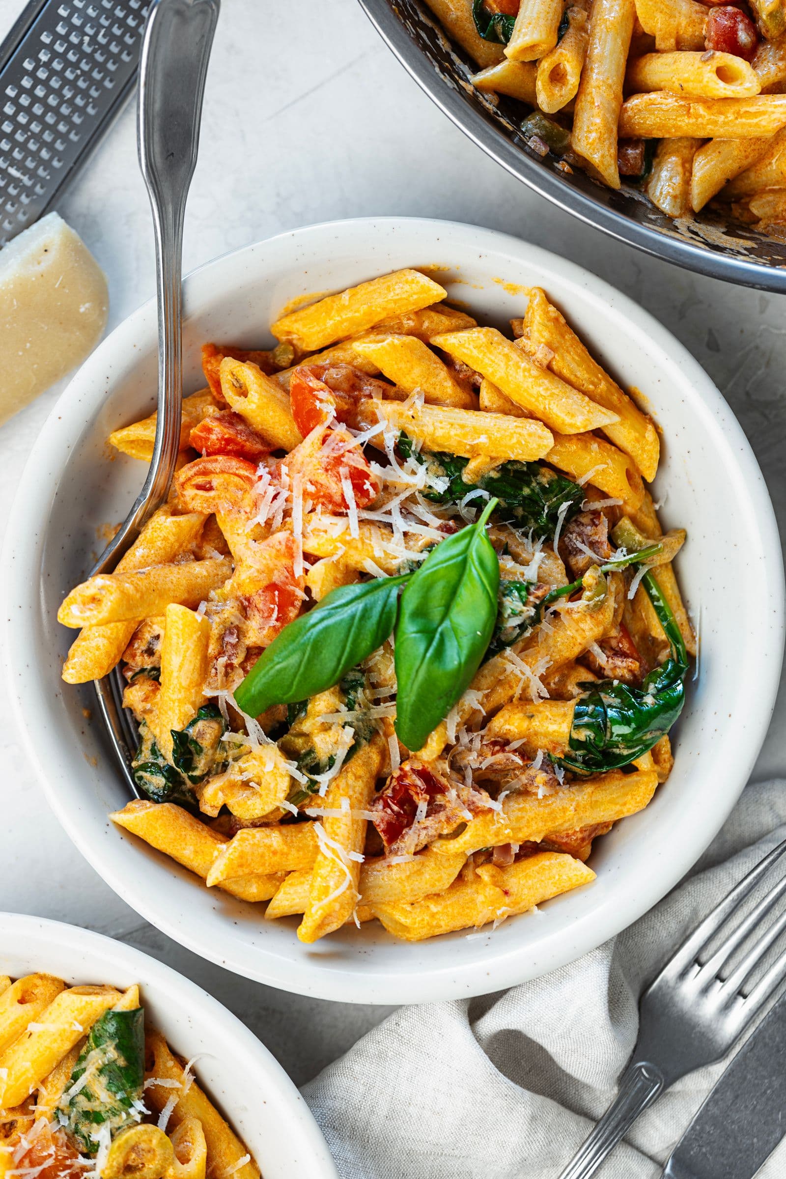 Vista de cima de uma taça de penne cremoso de tomate e espinafres ao lado da frigideira, com manjericão fresco