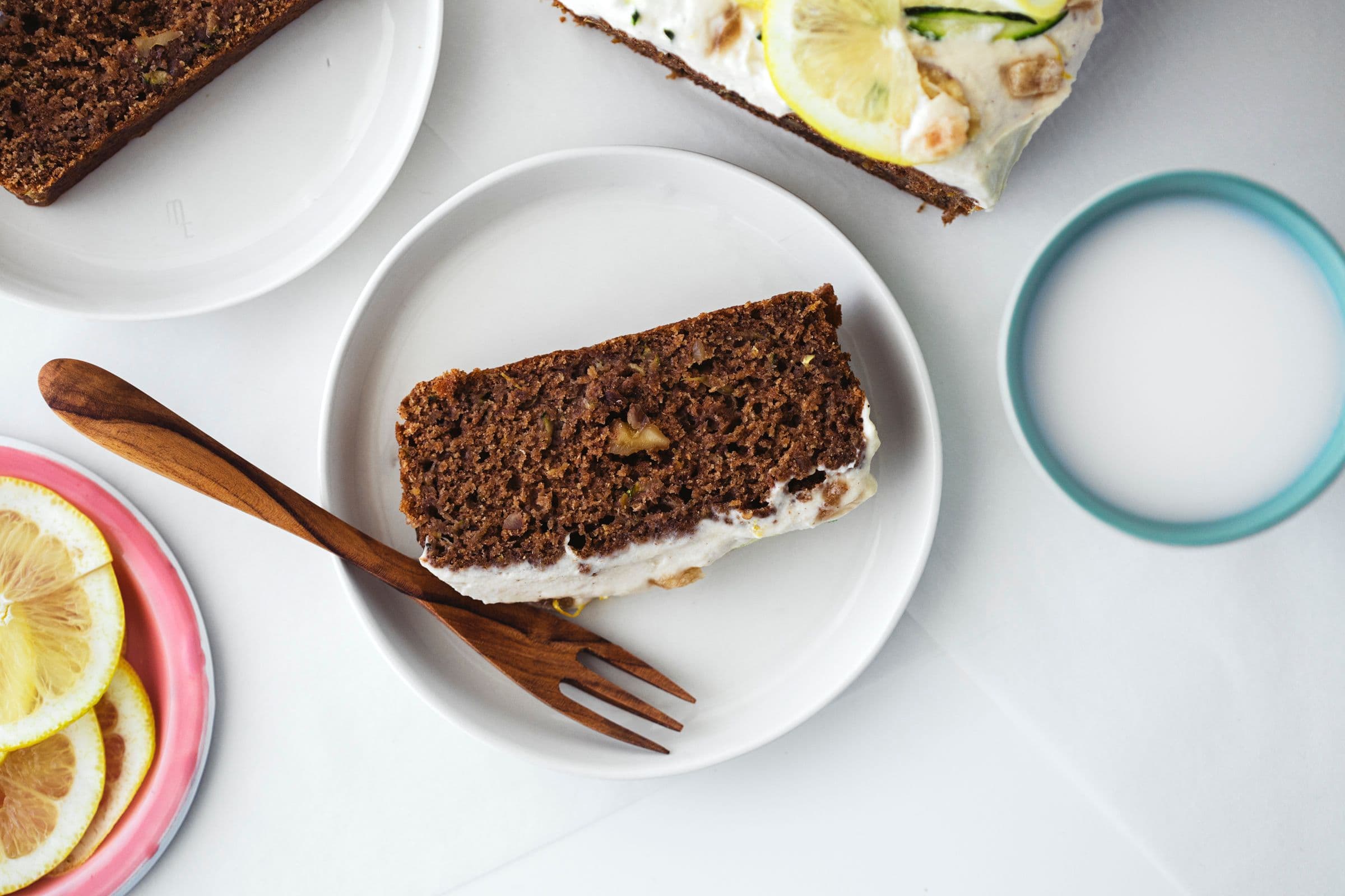 A slice of zucchini and lemon cake with frosting served on a white plate with a wooden fork.