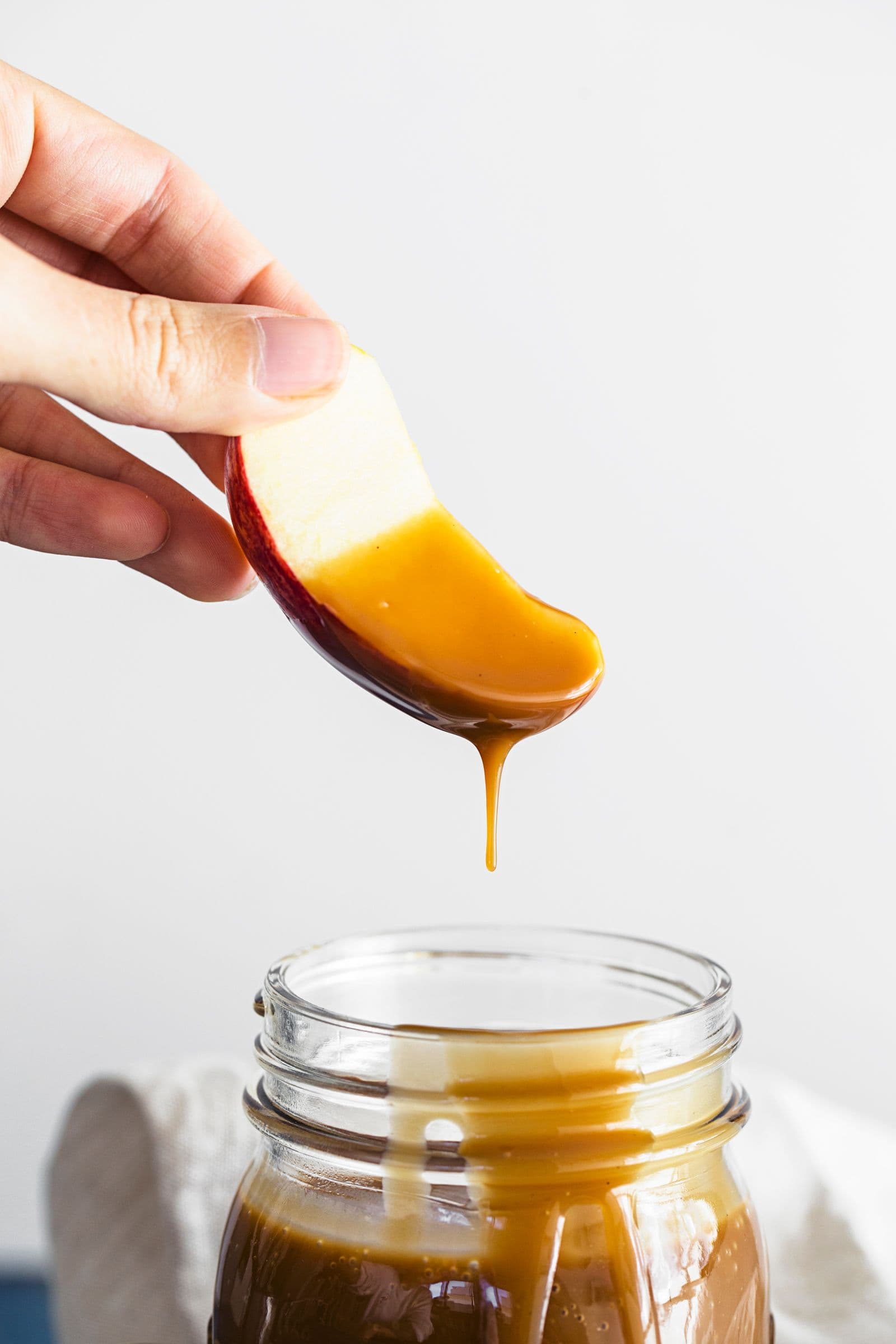 Hand holding a caramel-coated apple slice over a glass jar of sauce.