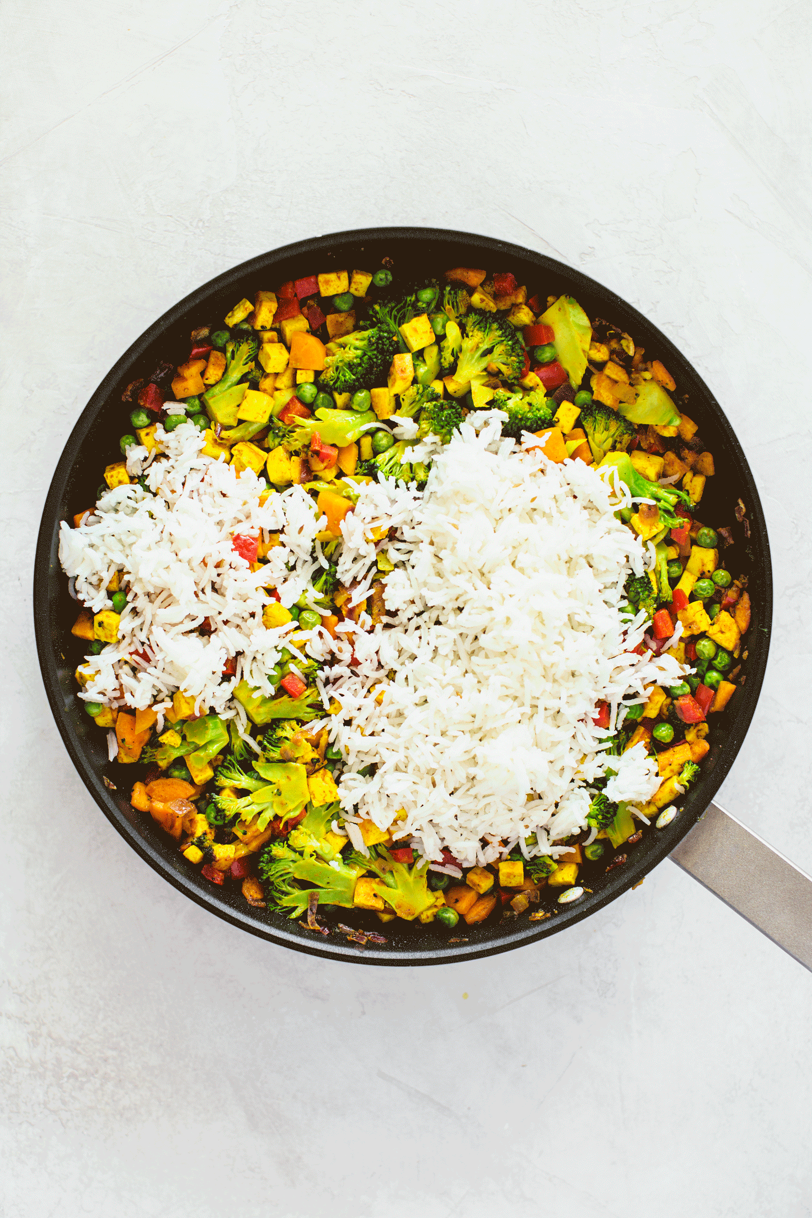 Animated gif of freshly cooked rice being added to sautéed vegetables in a black frying pan.