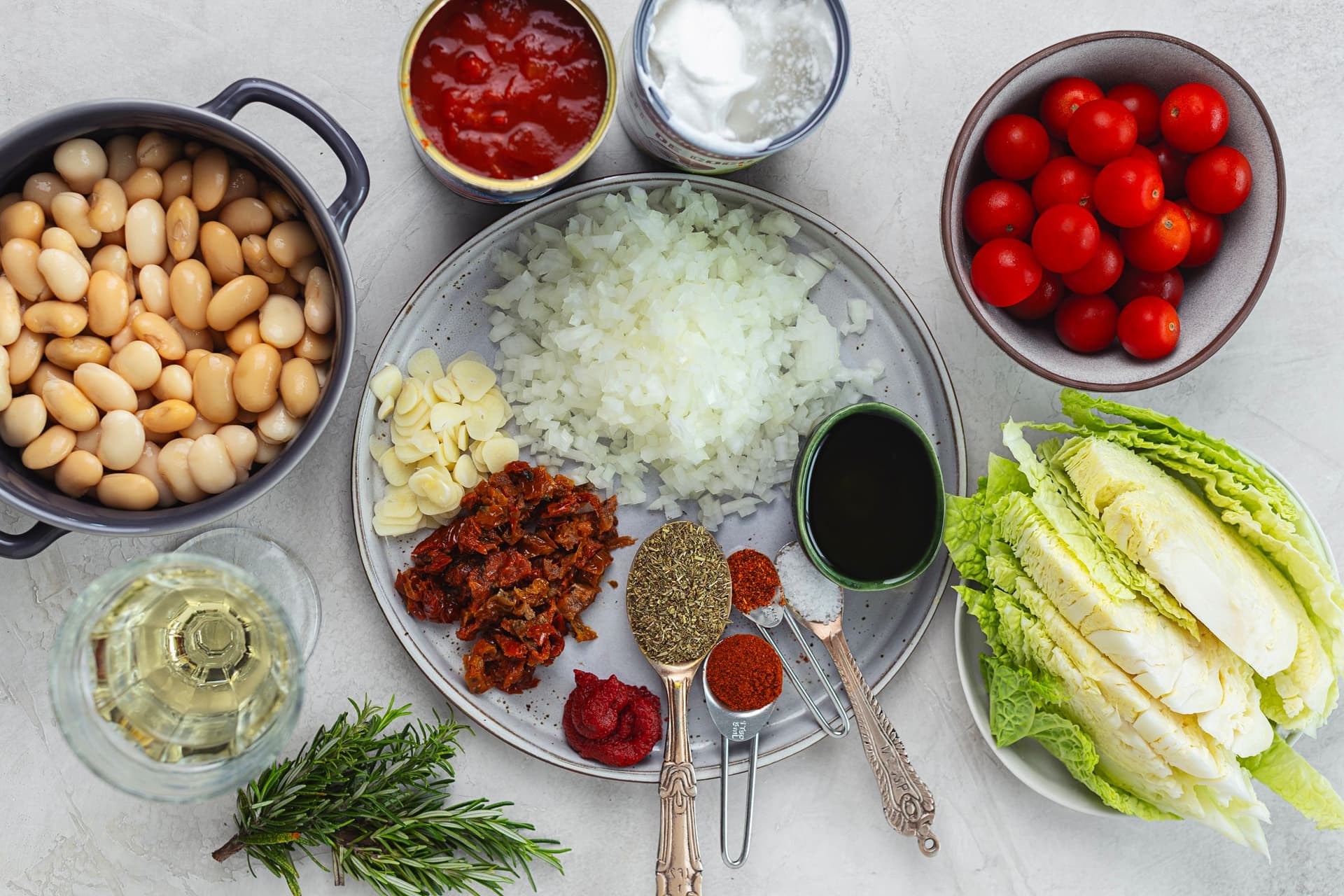 Vista de cima de todos os ingredientes do estufado dispostos sobre uma superfície clara: tomate-cereja, couve-lombarda, cebola picada, alho, tomate seco, especiarias, feijoca, tomate triturado, leite de coco e vinho branco.