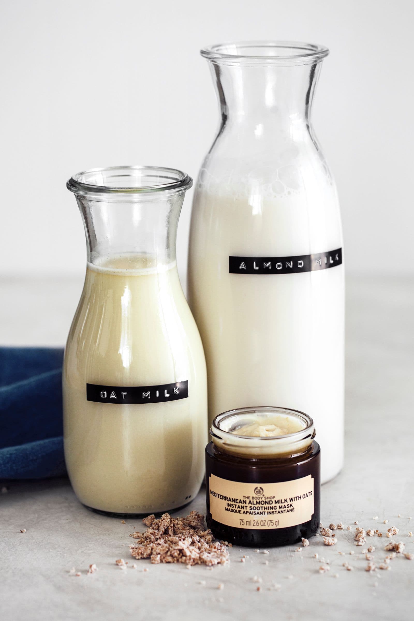 Garrafas de vidro com leite de aveia e leite de amêndoa, acompanhadas por um frasco aberto de máscara hidratante de amêndoa e aveia da The Body Shop.