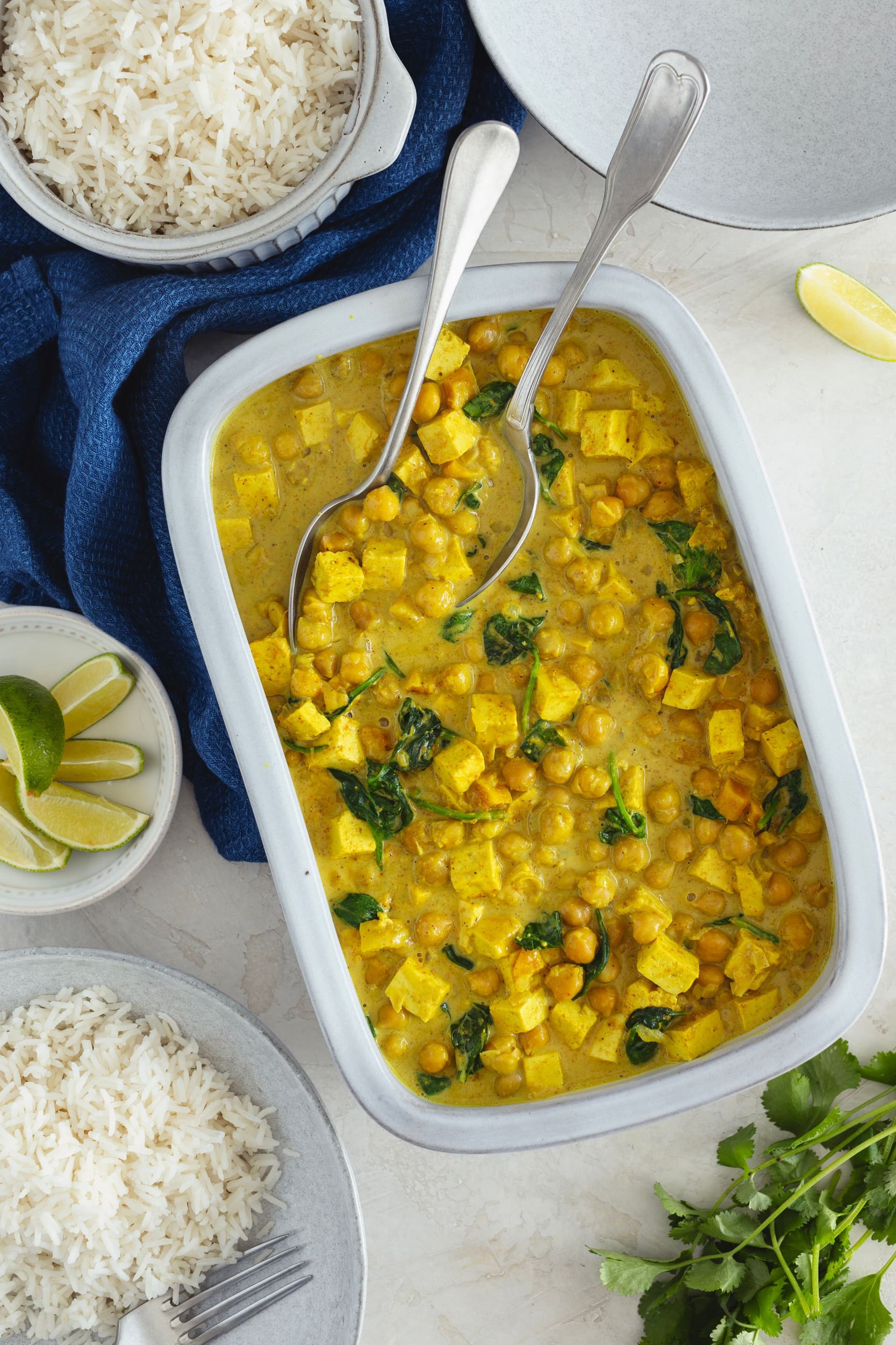 Large white baking dish filled with curry surrounded by bowls of white rice, lemon and herbs.