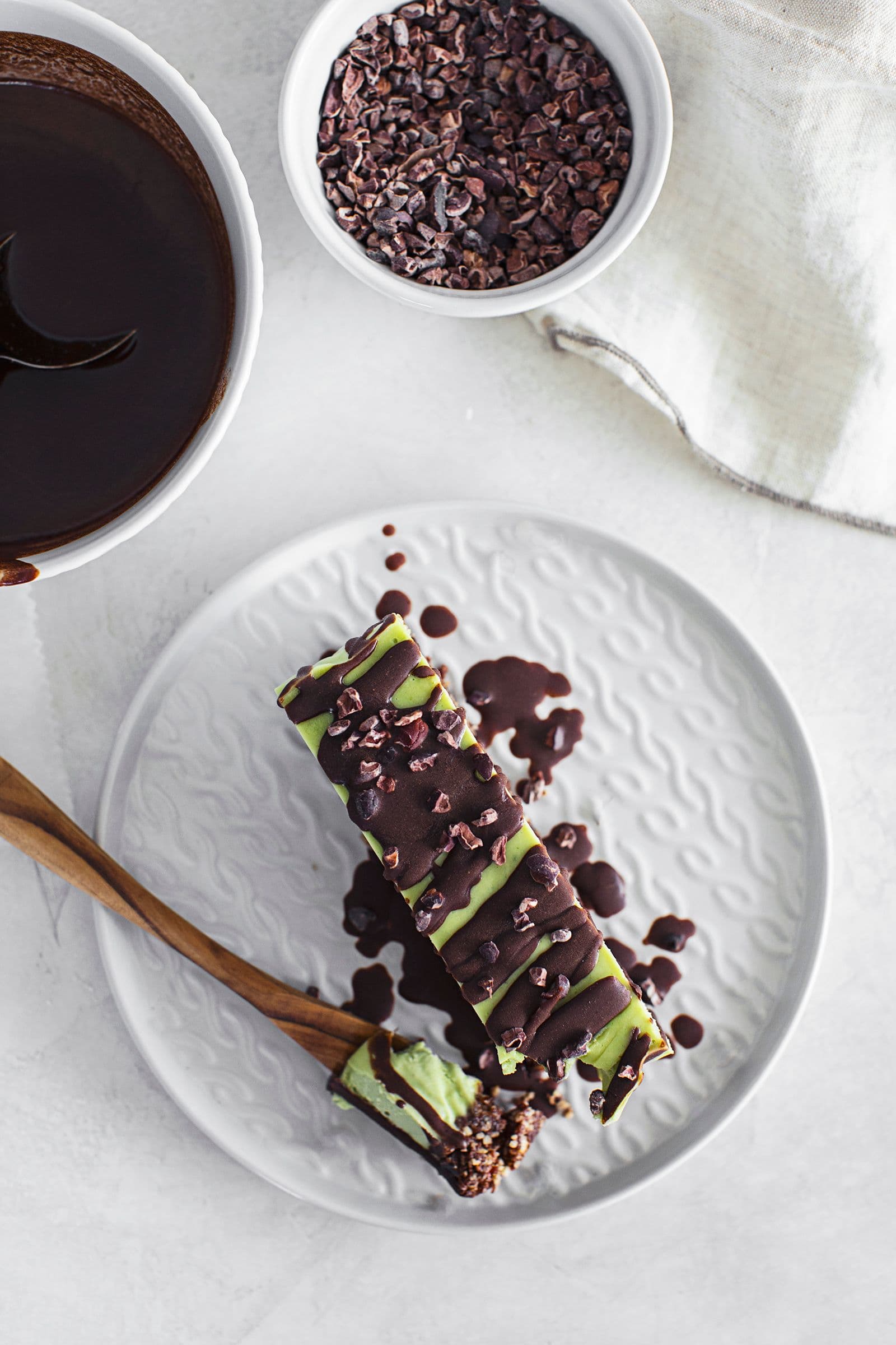 Fatia de cheesecake vegan de menta e chocolate com base de frutos secos, coberta com chocolate derretido e nibs de cacau.