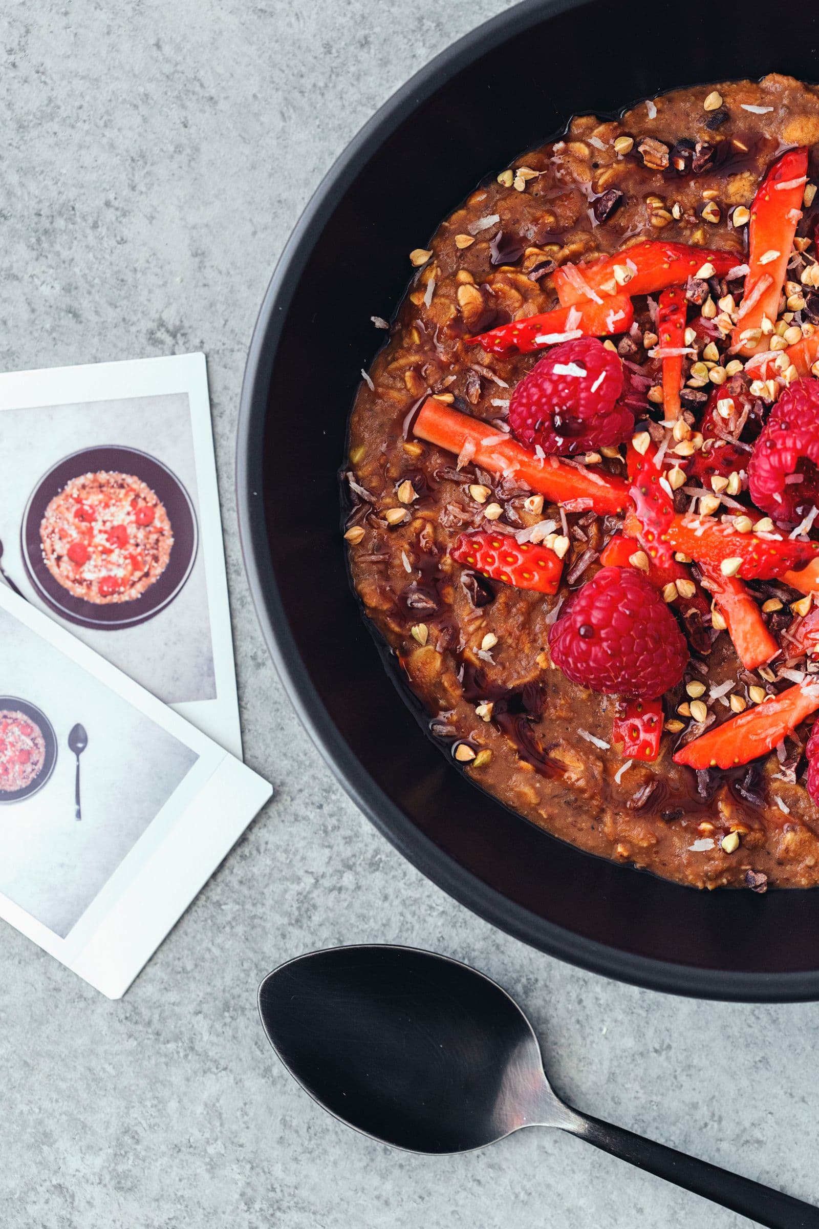 Bowl of cacao oatmeal with berries and seeds, next to a couple of mini polaroids and spoon.