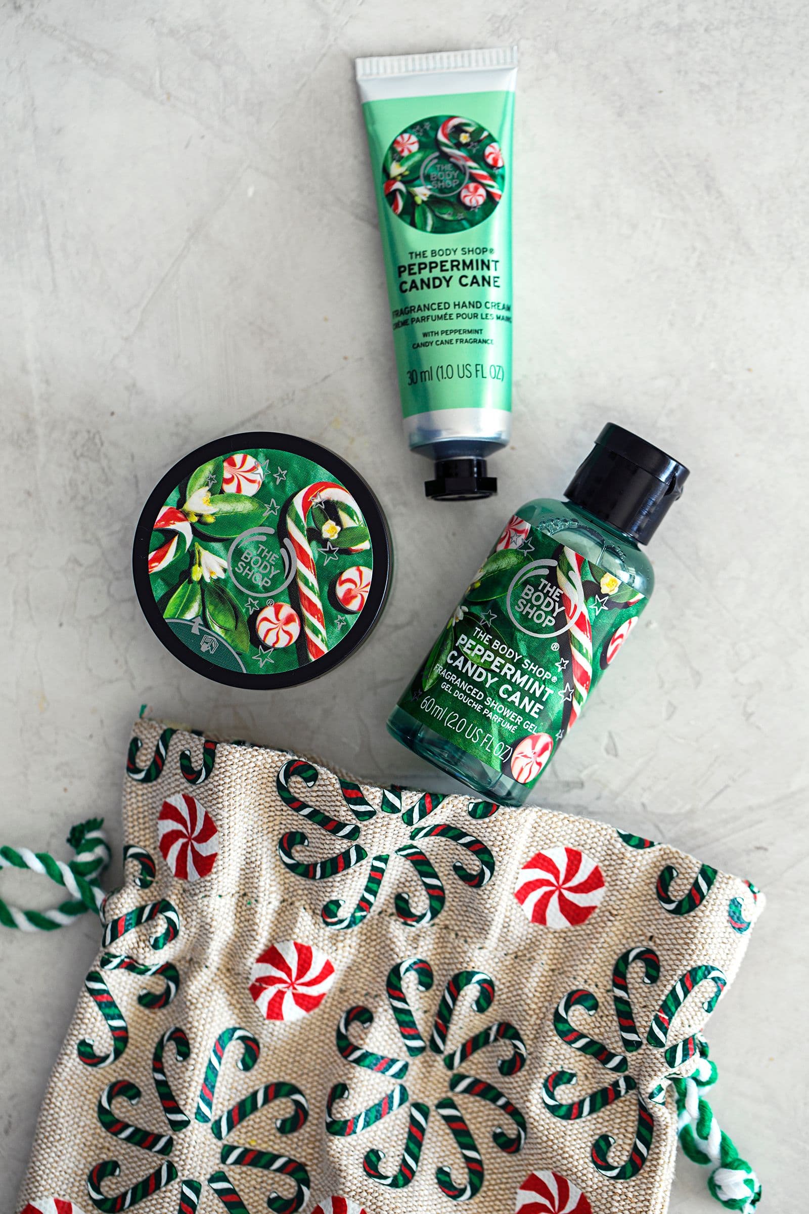 Conjunto de produtos The Body Shop "Peppermint Candy Cane" sobre superfície clara, ao lado de um saco de tecido com padrão natalício.