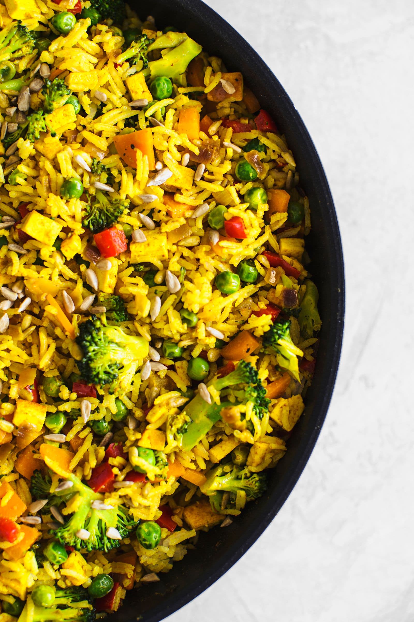 Vista aproximada de frigideira com arroz frito vegan colorido com tofu, brócolos e legumes sobre superfície branca.