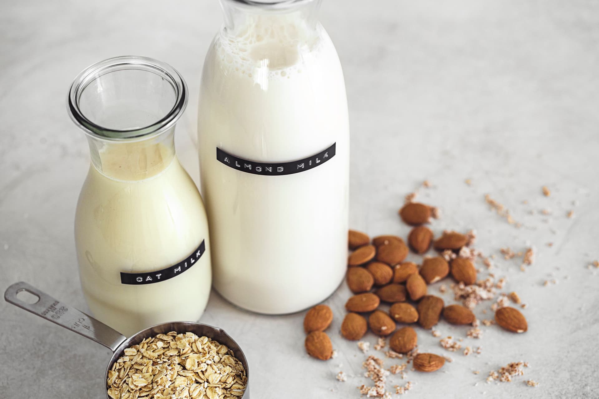 Ingredientes básicos para fazer leite vegetal, com frascos de leite de aveia e de amêndoa, aveia em flocos e amêndoas inteiras.