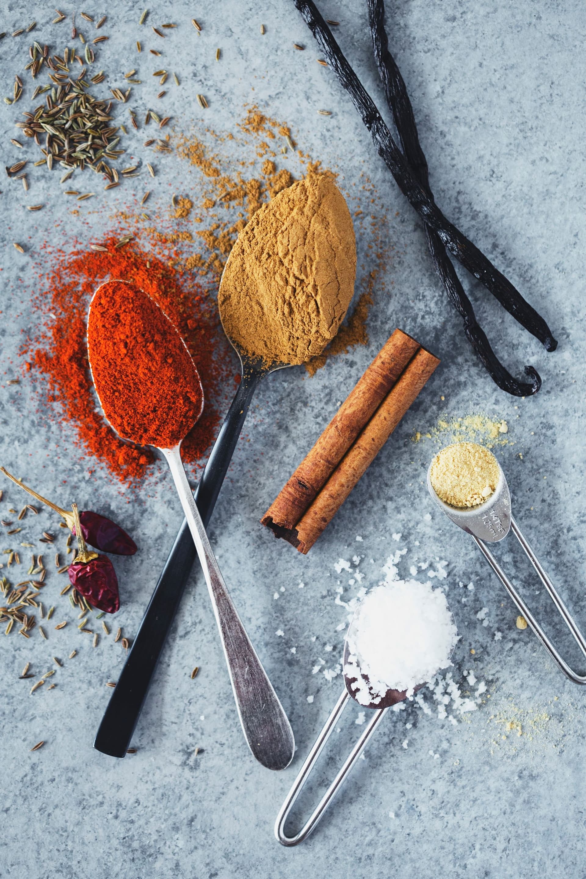 Vista de cima de especiarias como paprika, malagueta, cominhos, baunilha, canela, alho e sal com colheres medidoras.