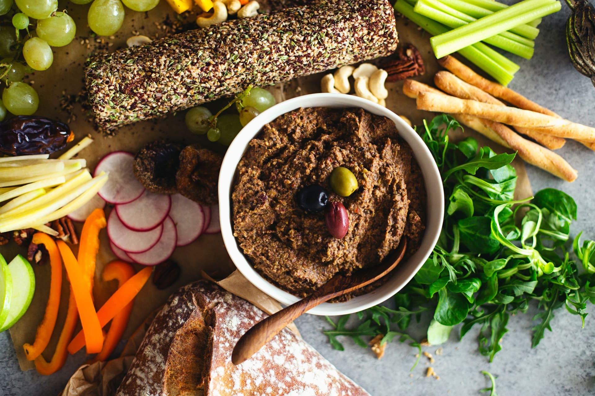 Rolo de queijo de caju e paté de lentilhas servidos numa travessa grande com uvas, frutos secos, legumes crus e fatias de pão.