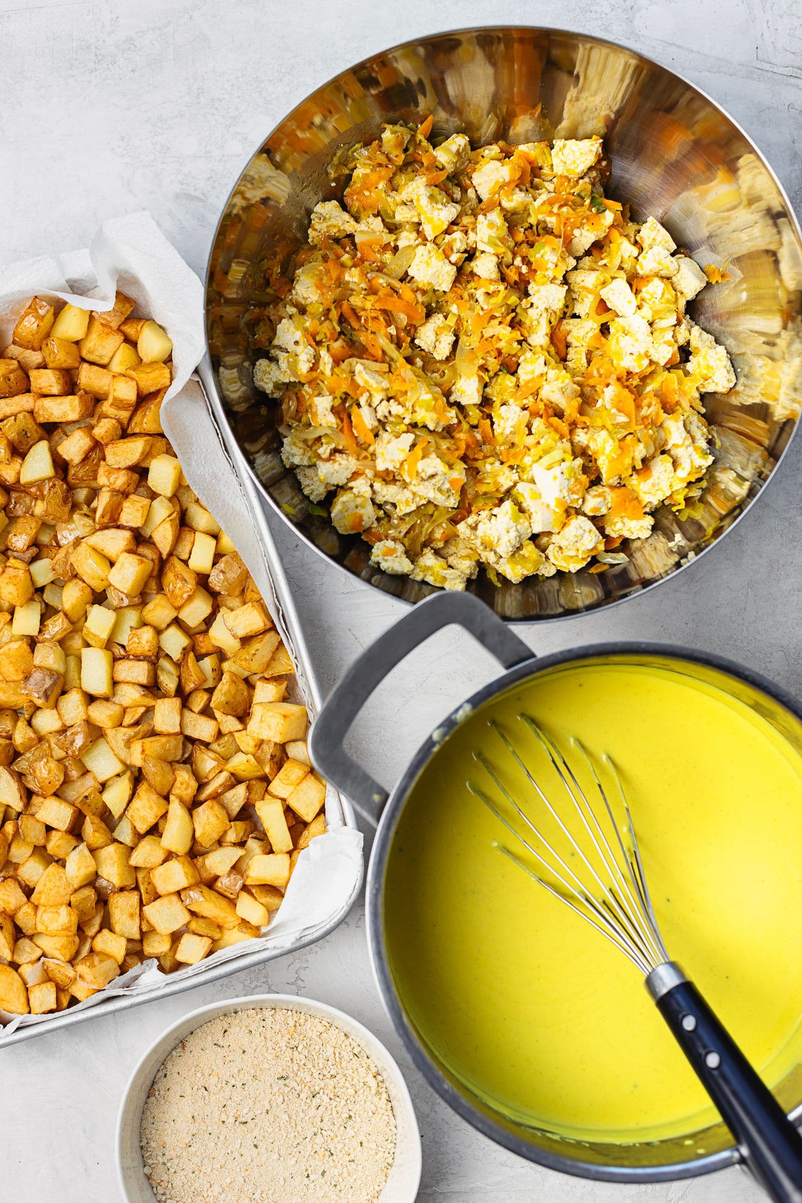 Refogado de tofu e legumes numa taça, batatas fritas douradas e molho béchamel pronto a usar.