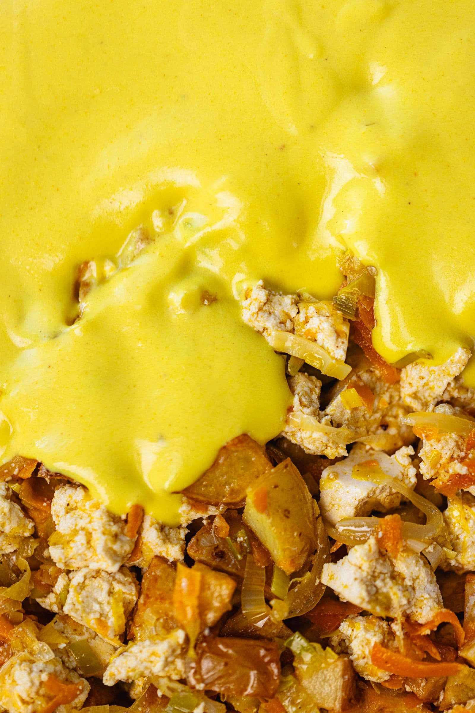 Close-up do béchamel cremoso a espalhar-se sobre o tofu, batatas e legumes.