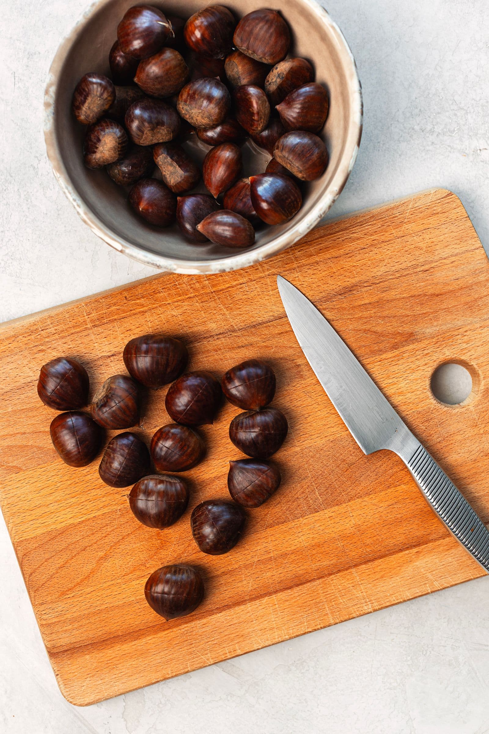 “Wooden board with raw scored chestnuts and a knife, with a bowl of whole chestnuts on the side