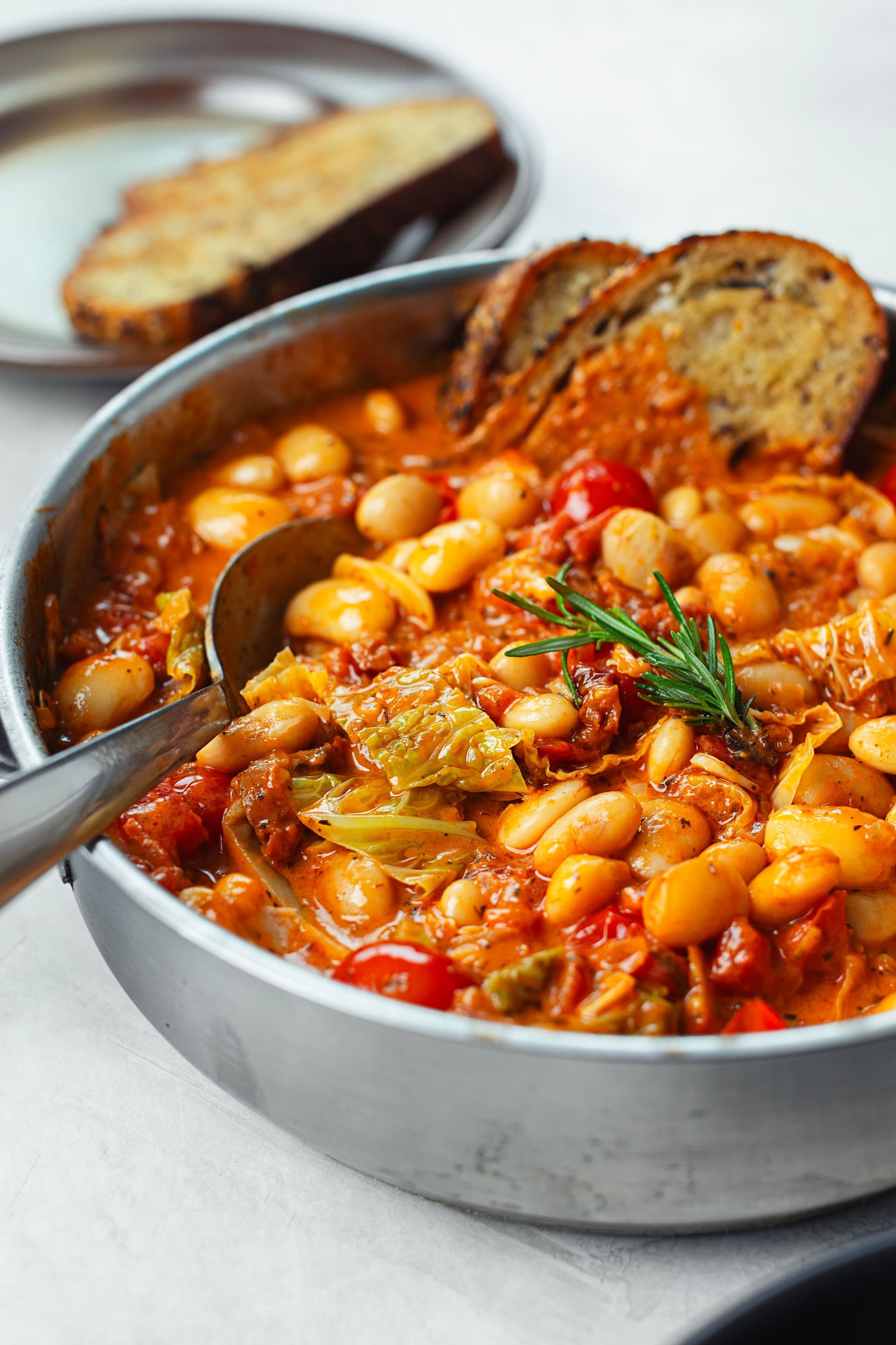Vista lateral da panela cheia do estufado cremoso de tomate e coco com feijoca, com fatias de pão torrado encostadas ao interior.