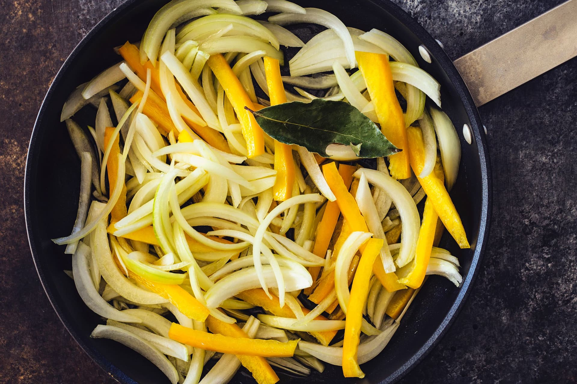 Frigideira preta com cebolas, pimentos amarelos e uma folha de louro a saltear.