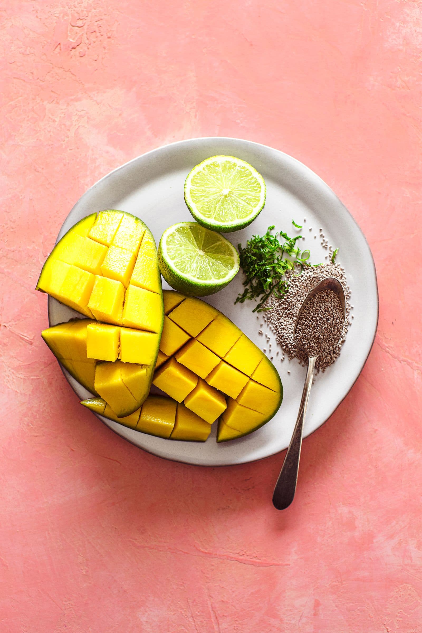Plate with diced mango, lime halves, lime zest and chia seeds on a pink surface.