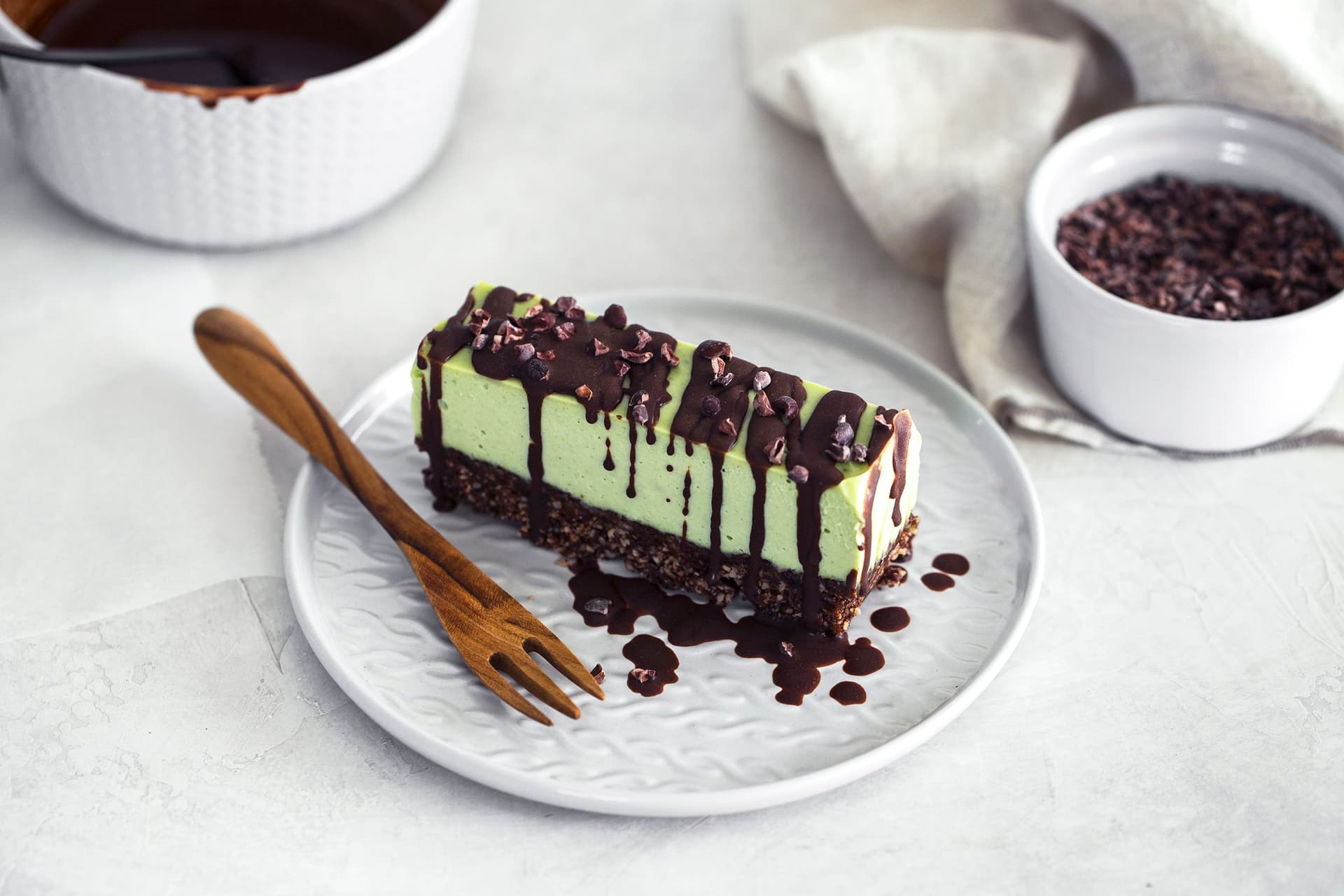 Fatia de cheesecake vegan de menta e chocolate com base de frutos secos, coberta com chocolate derretido e nibs de cacau.