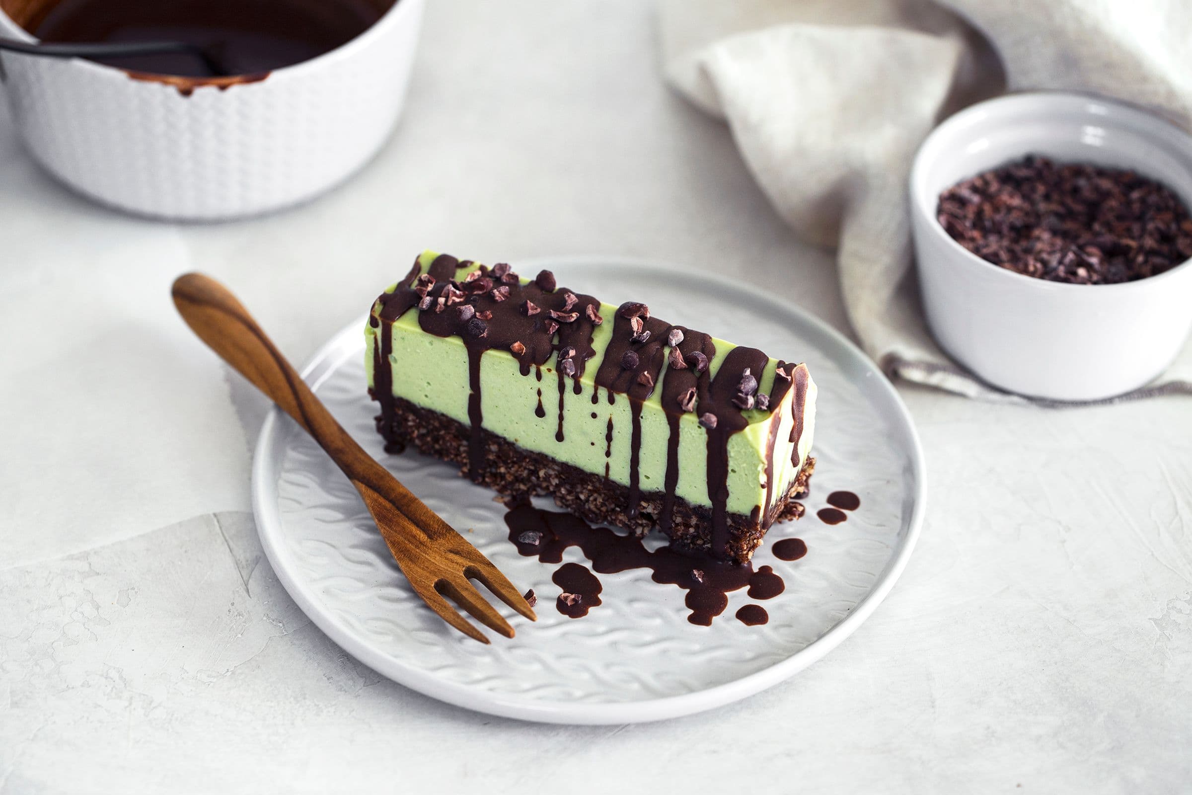 Fatia de cheesecake vegan de menta e chocolate com base de frutos secos, coberta com chocolate derretido e nibs de cacau.