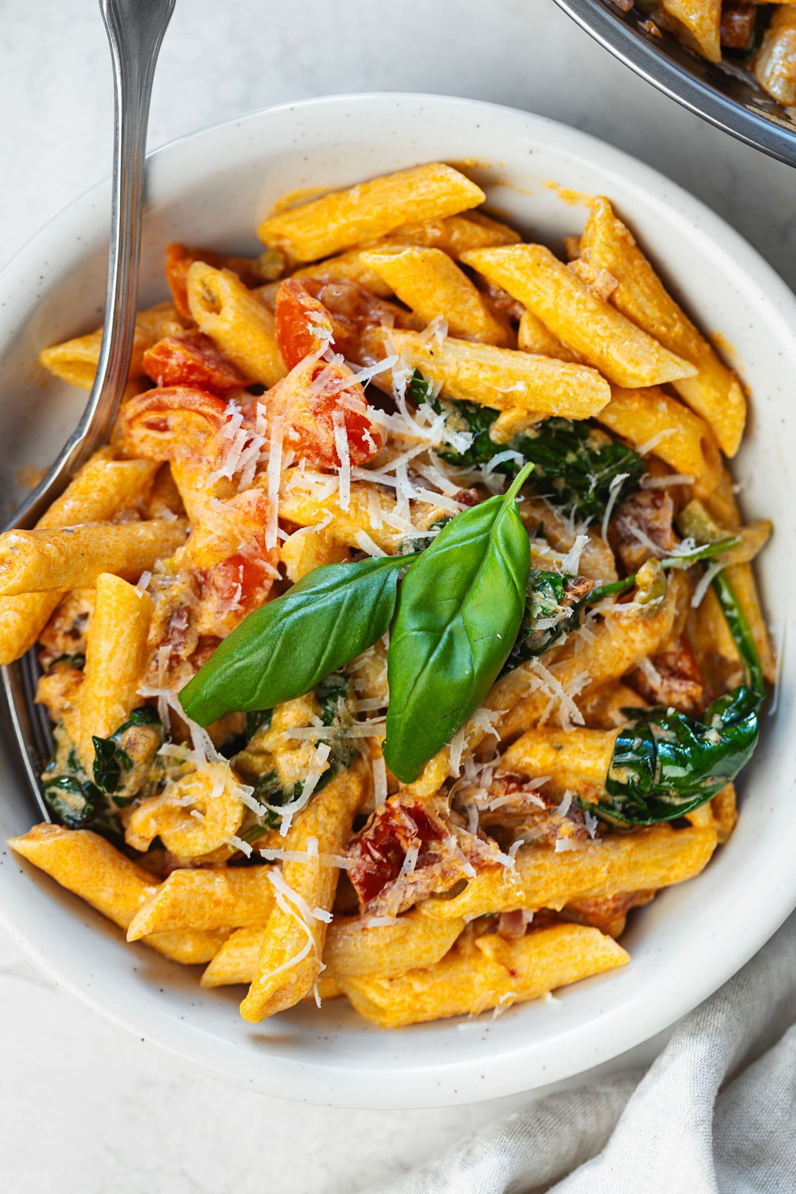 Close-up of a bowl of creamy tomato and spinach penne topped with fresh basil and grated vegan parmesan