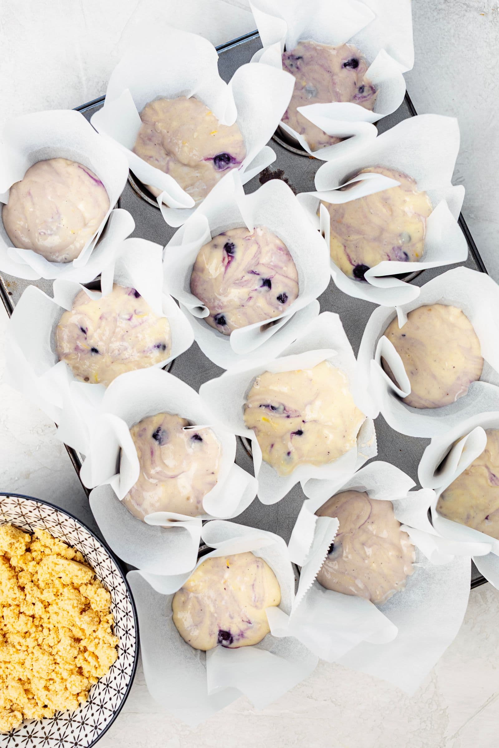 Massa de queques de limão e mirtilos distribuída numa forma de muffins forrada com papel vegetal.