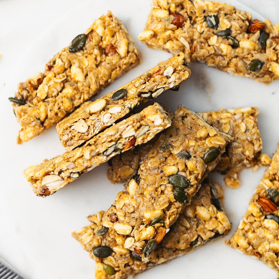 Barras proteicas caseiras com aveia, sementes, frutos secos e tahini, cortadas e empilhadas num prato de mármore.
