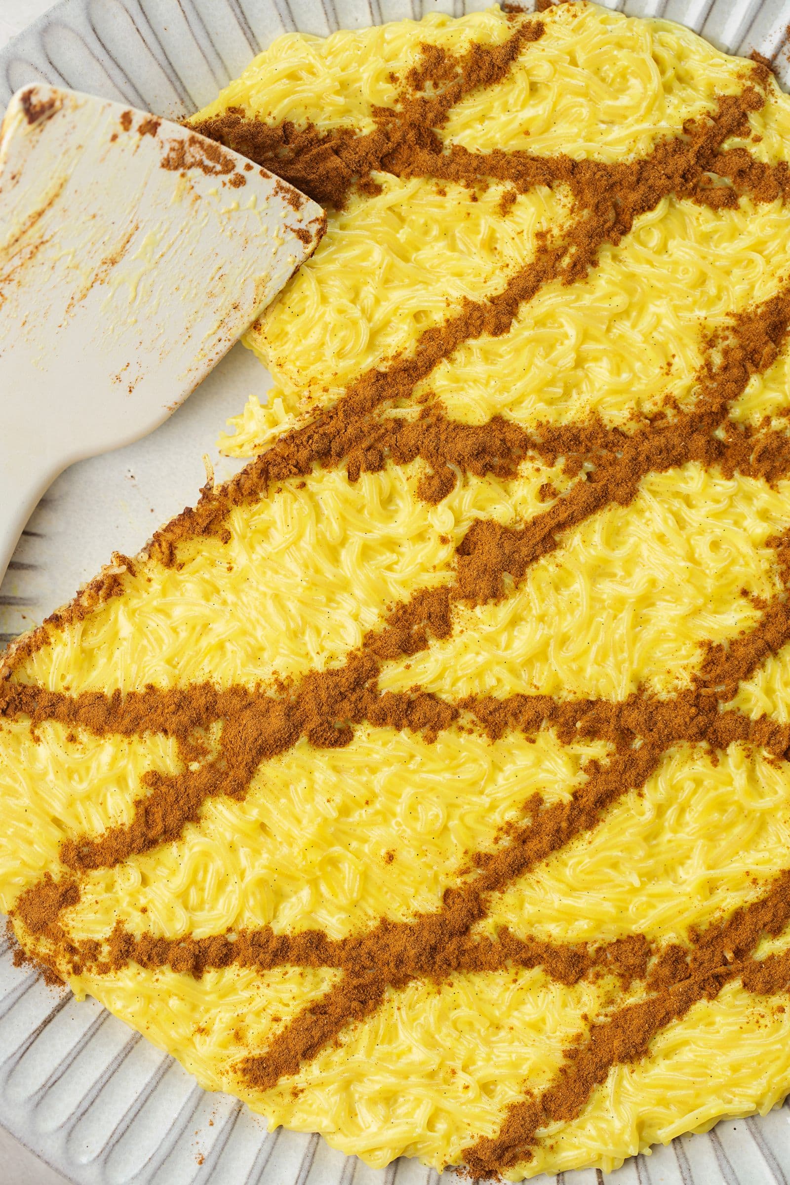 Detalhe da aletria vegan num prato de servir com uma espátula, a mostrar a textura cremosa e o padrão de canela por cima.