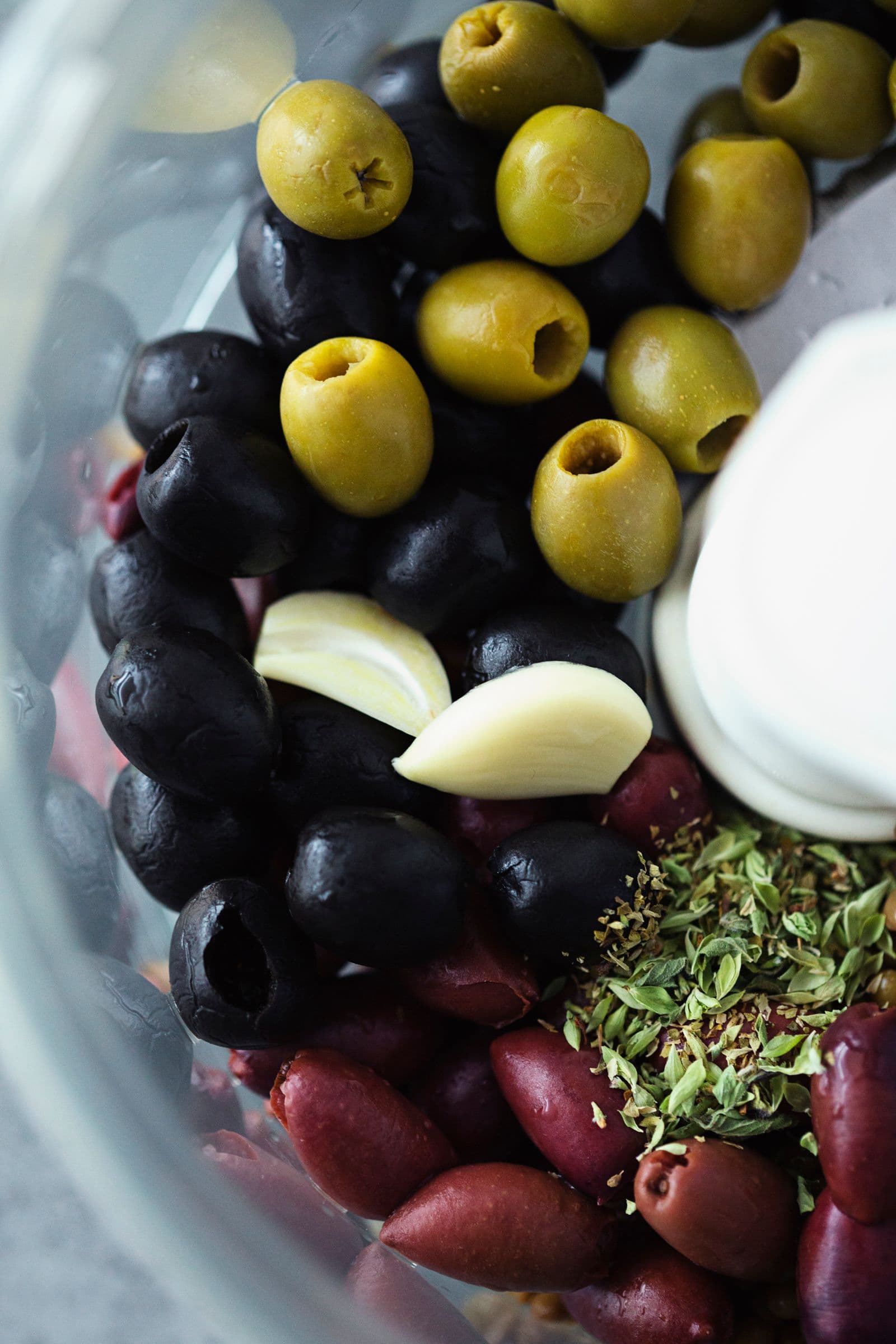 Assortment of black and green olives with garlic cloves and herbs in a food processor bowl.