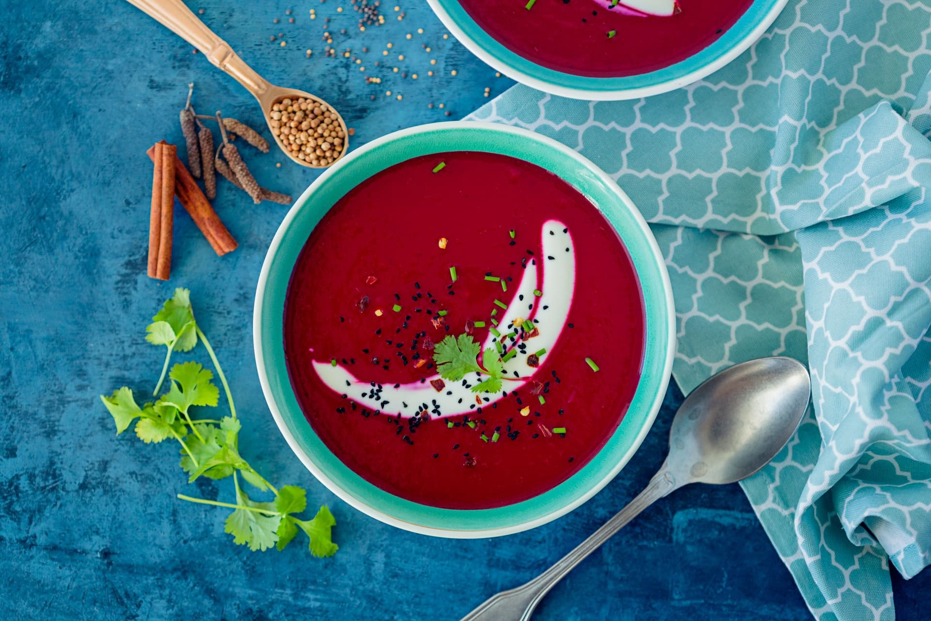Vibrant beetroot and reinette apple soup served in a turquoise bowl, topped with coconut yogurt and fresh herbs on a blue background.
