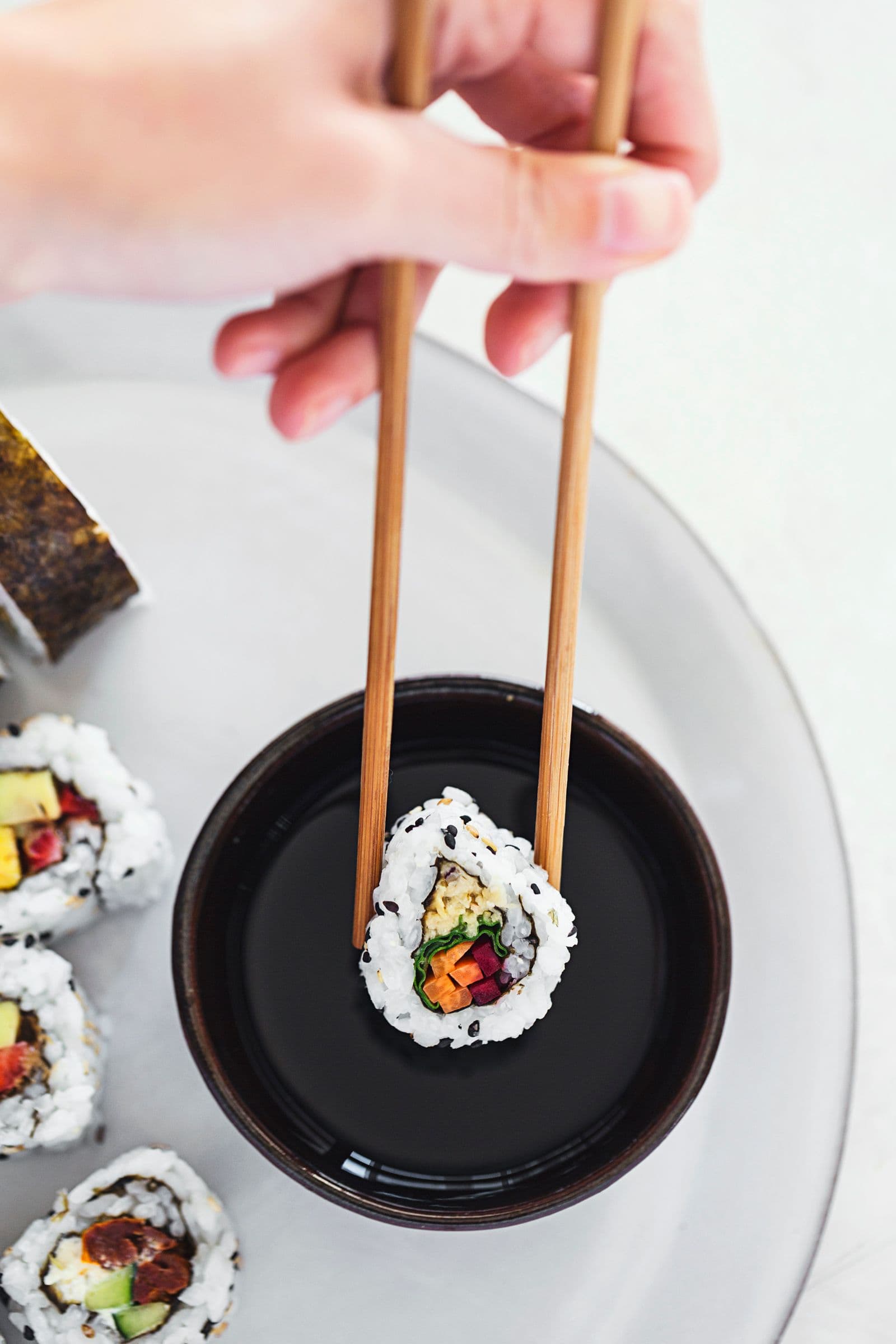 Close-up of a hand dipping a vegan sushi roll into soy sauce using chopsticks.