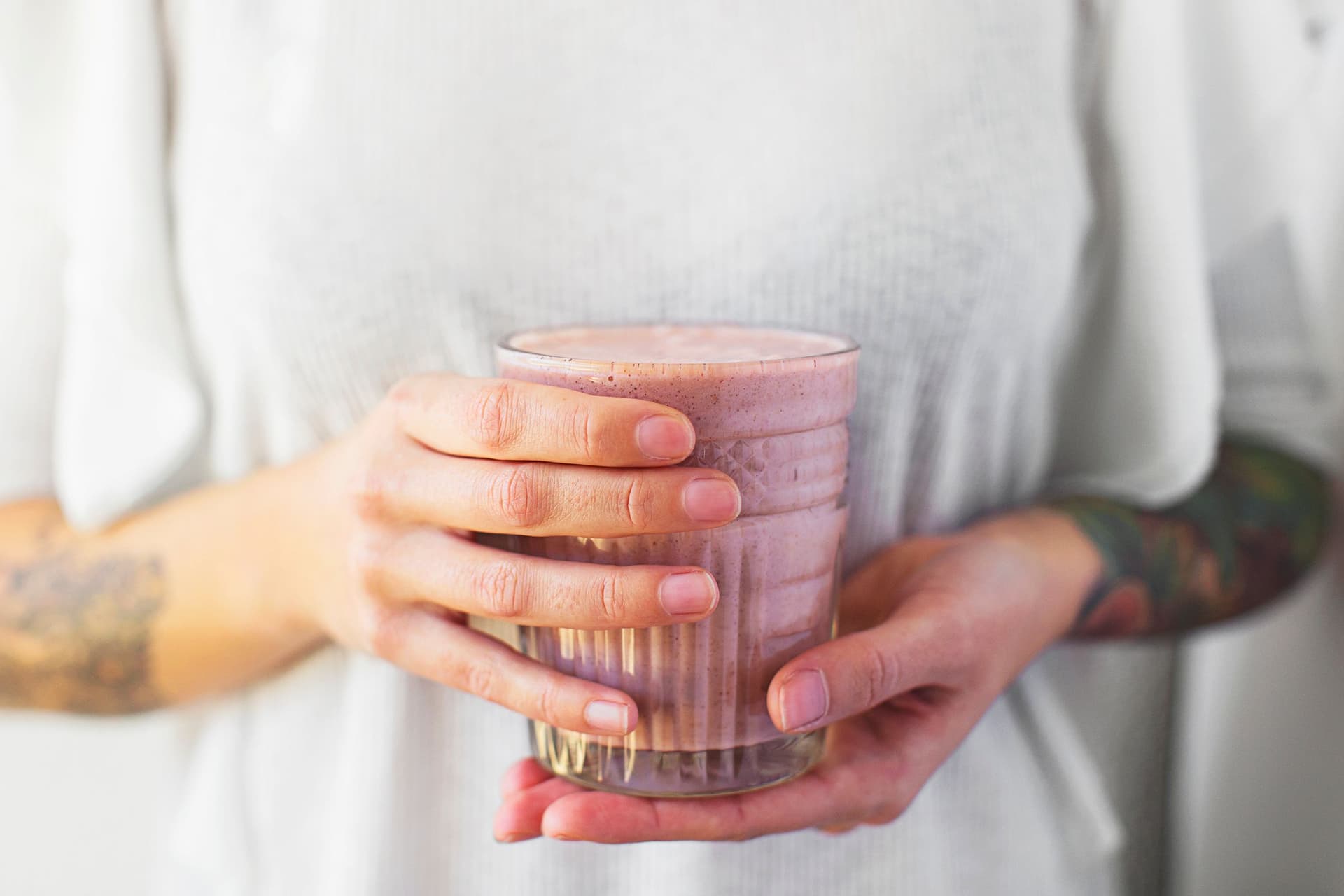 Person holding a glass of a light pink smoothie with both hands.