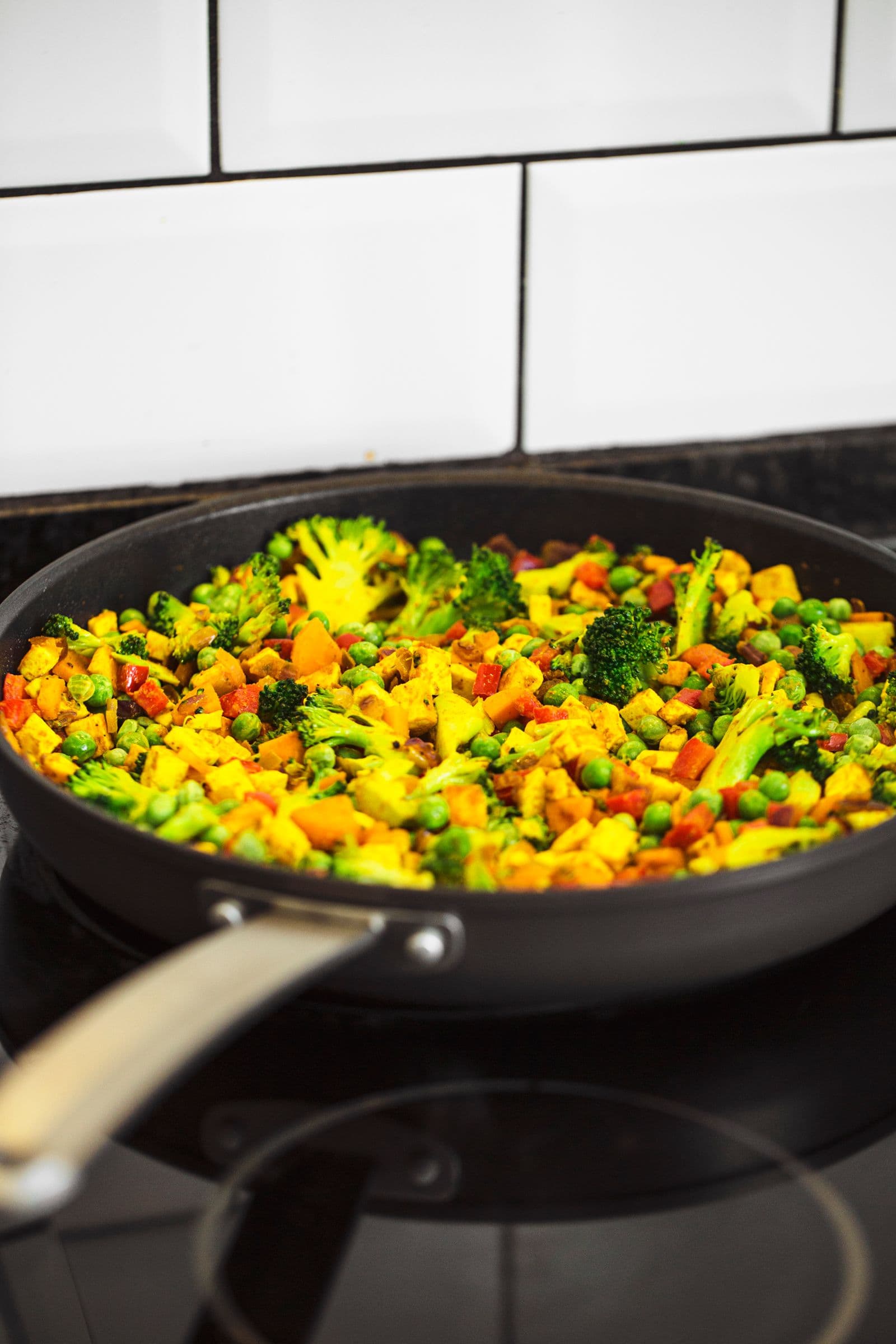 Frying pan on a stovetop with carrots, broccoli and peppers being sautéed.