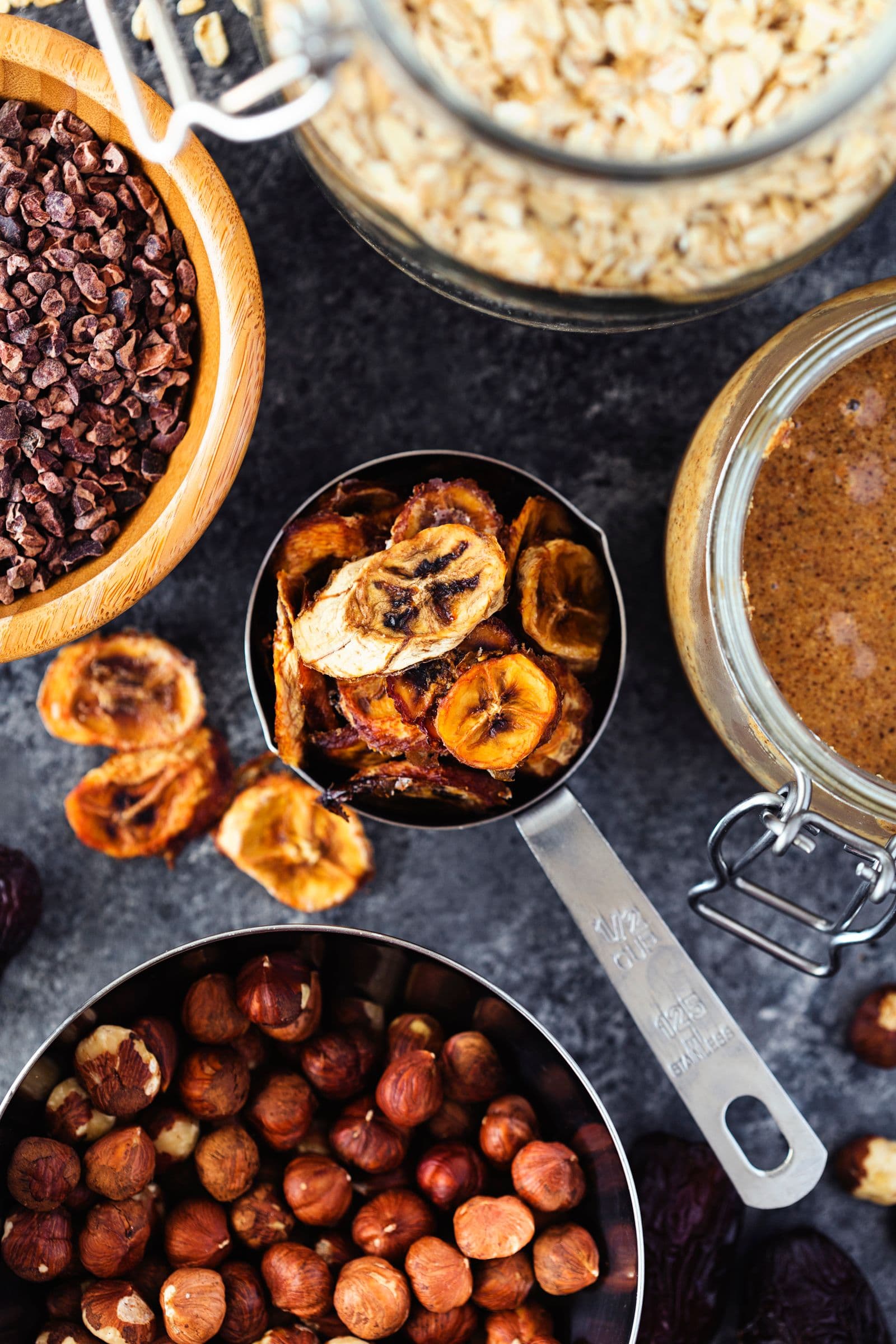 Ingredientes variados, incluindo tâmaras, avelãs, chips de banana e uma taça com manteiga de oleaginosas.