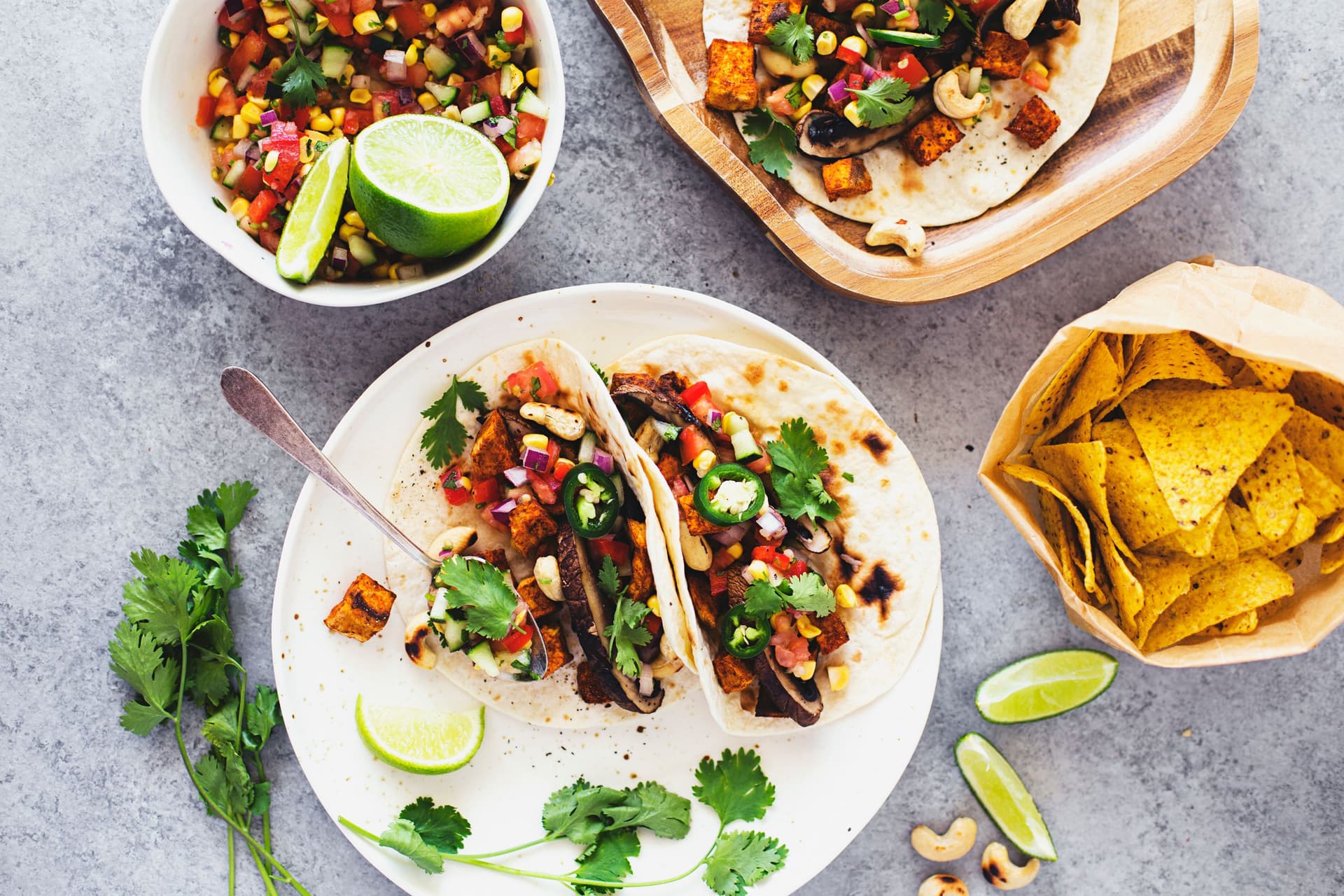 Prato de tacos com cogumelos grelhados, batata-doce temperada, salada e ervas frescas, servidos com gomos de lima e nachos.