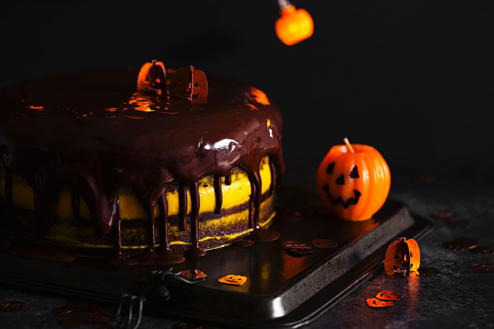 Halloween layered cake with dripping chocolate glaze, topped with tiny pumpkin decorations and orange lights in the background.
