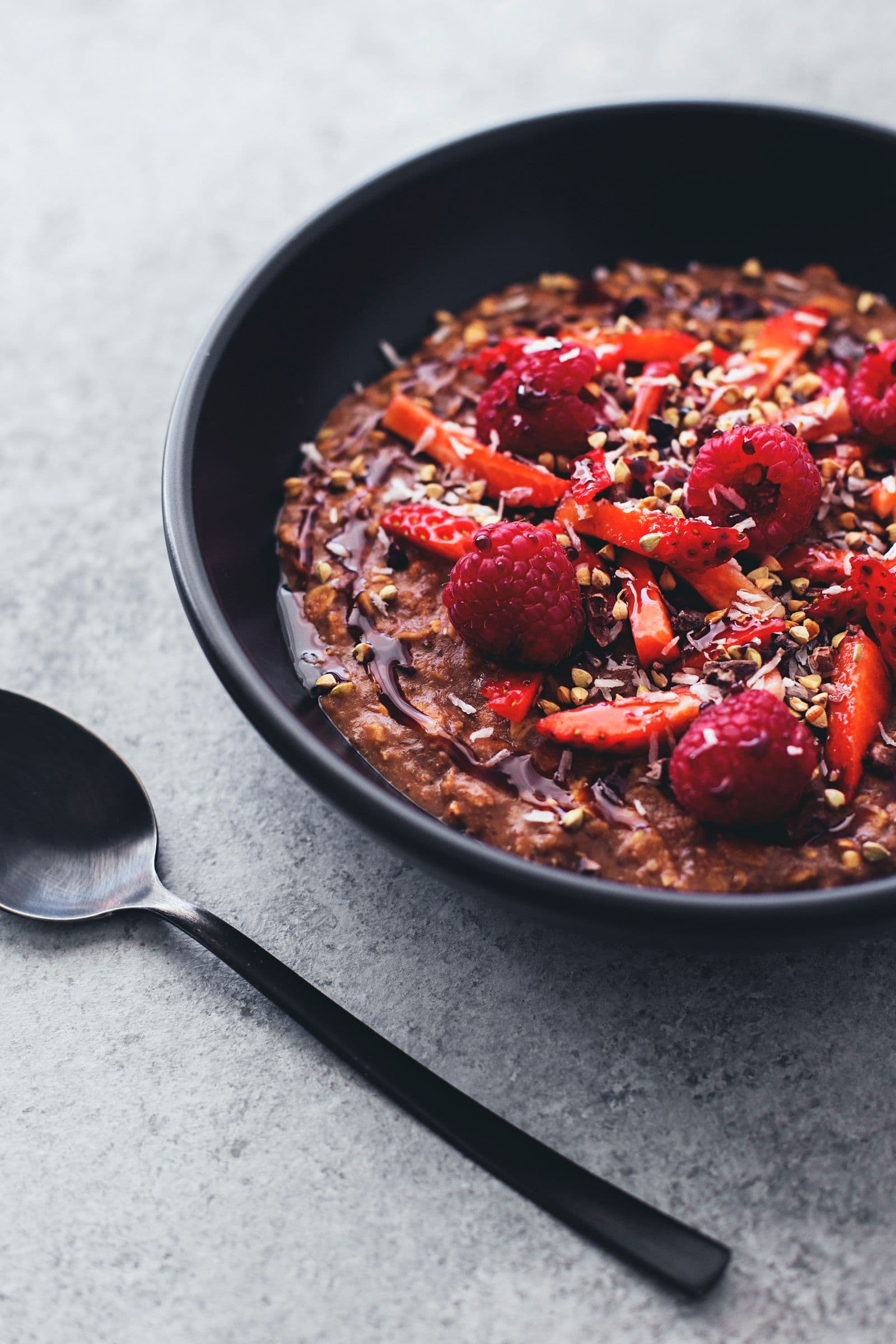 Black bowl of cacao oatmeal topped with berries and seeds, with a black spoon on the side.