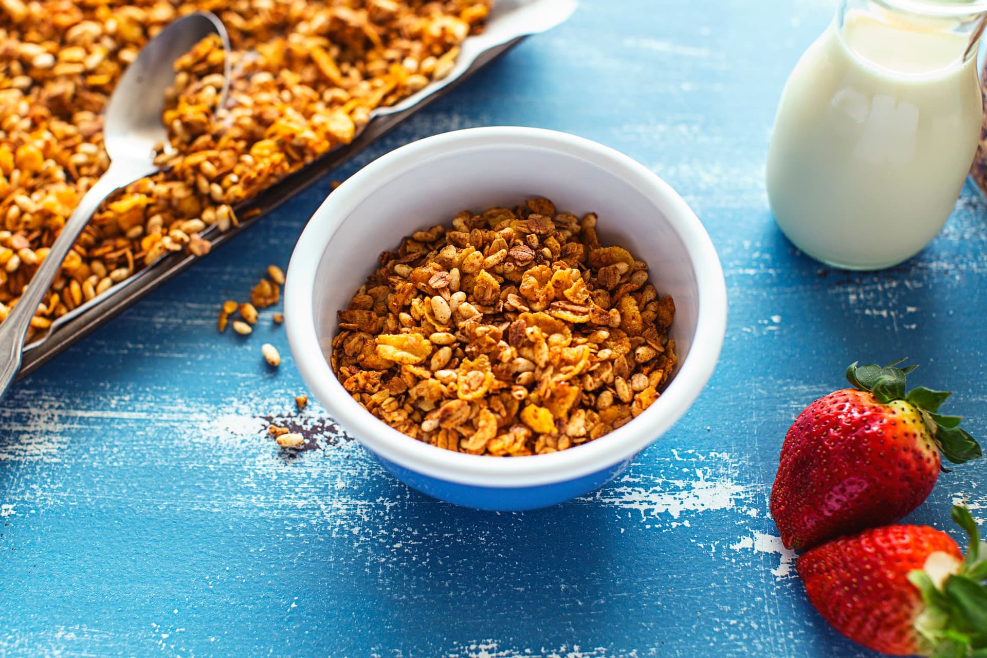 Uma taça de granola super crocante com cornflakes, servida com morangos frescos e bebida vegetal sobre uma mesa azul.