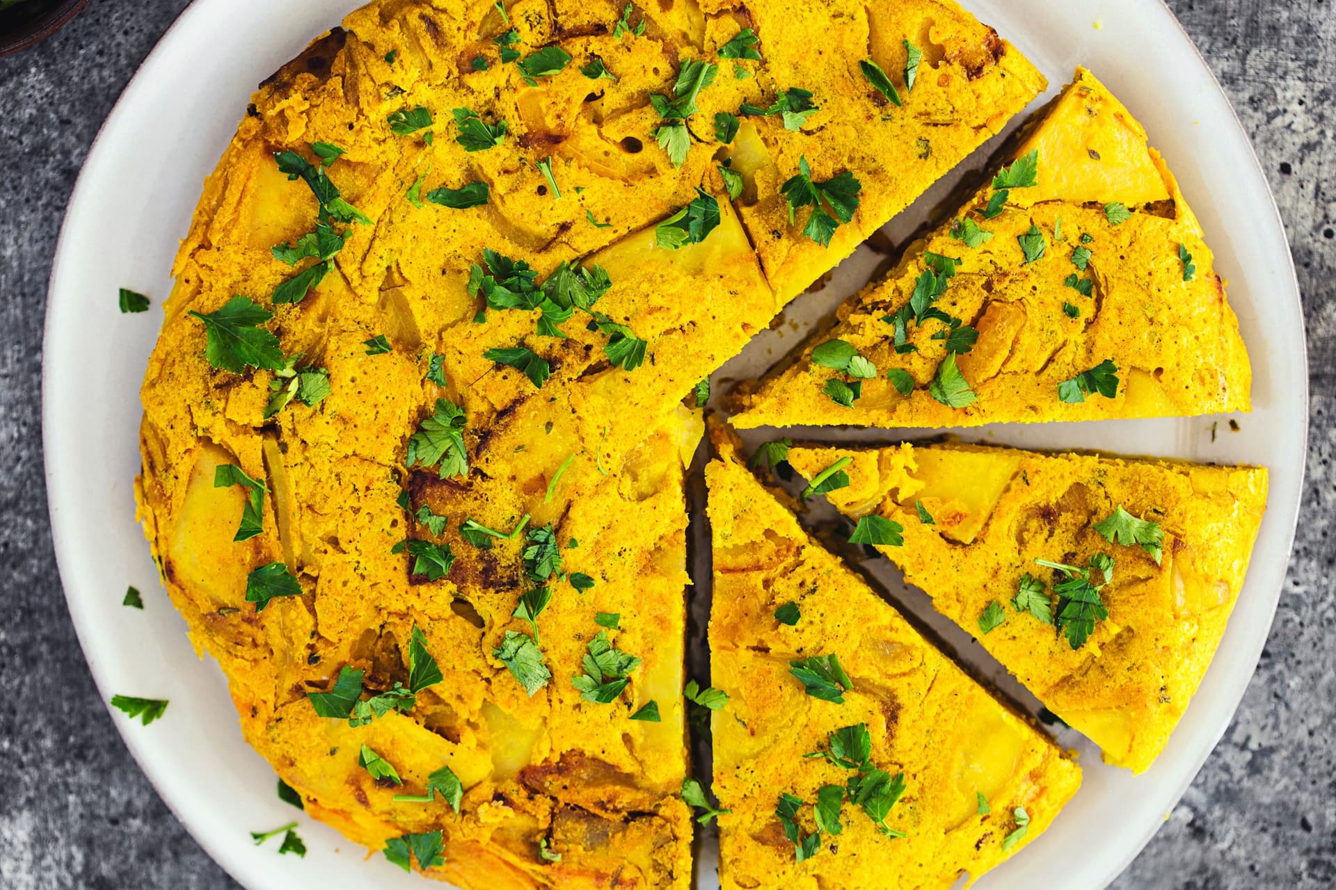 Tortilha espanhola vegan num prato branco, decorada com salsa, com três fatias cortadas.