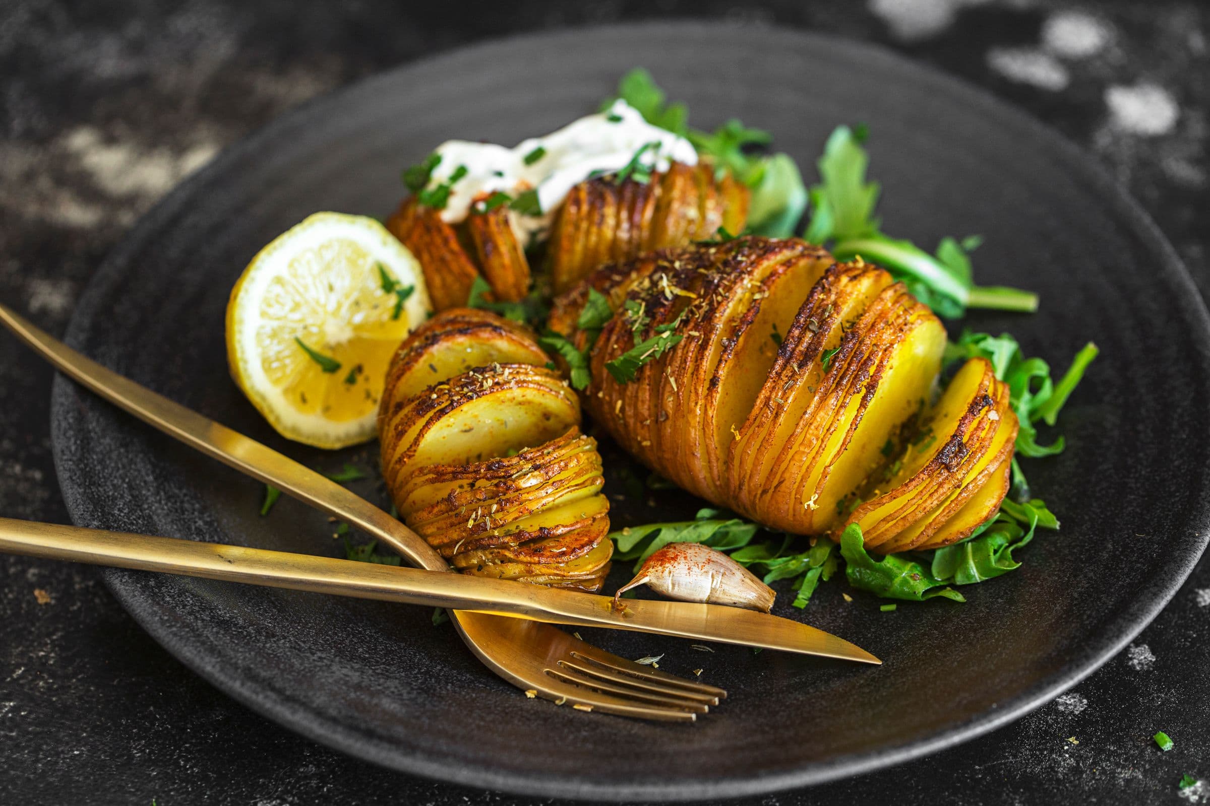 Golden Hasselback potatoes with herbs, spices and creamy sauce, served with fresh arugula on a black plate.