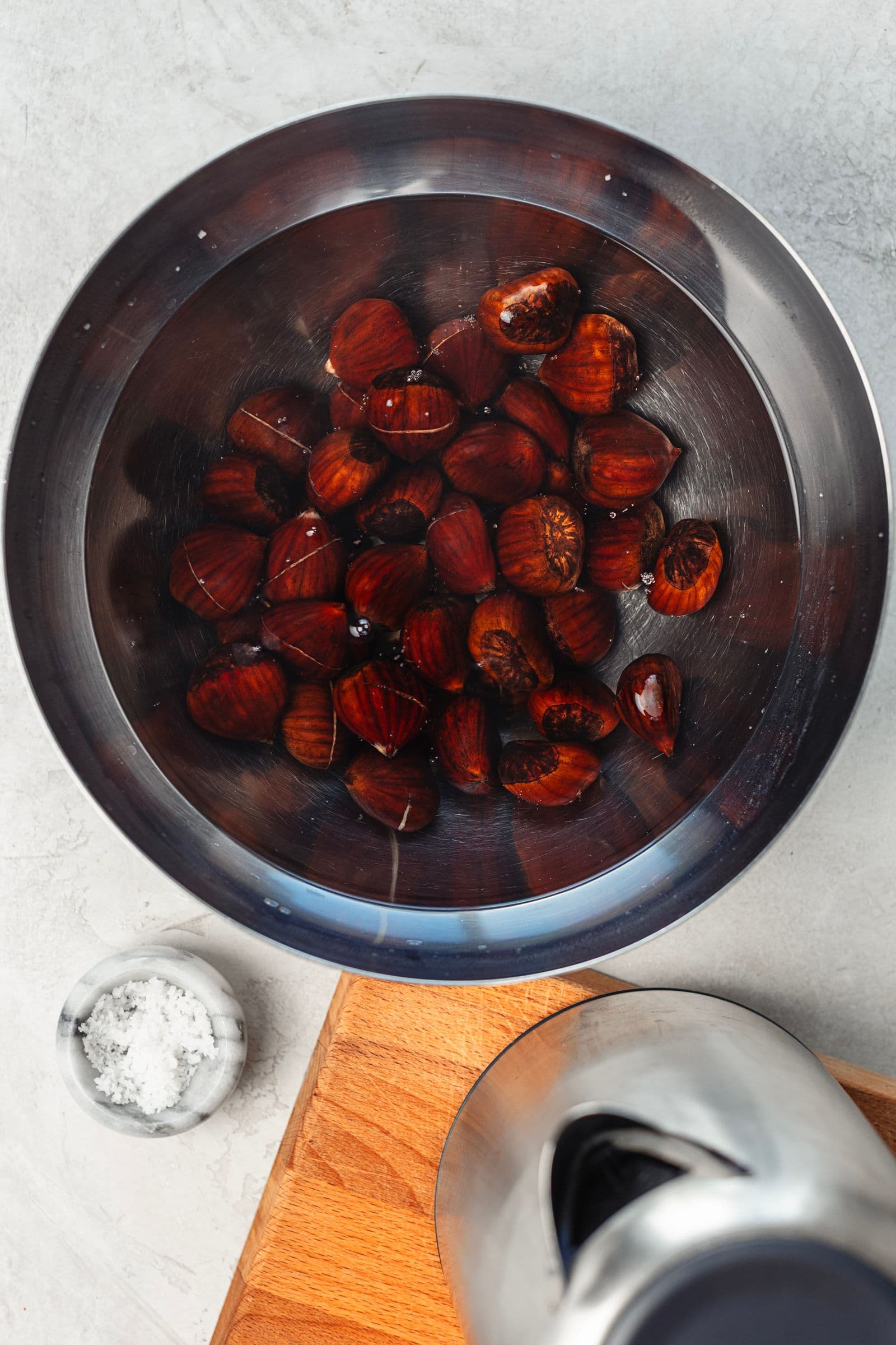Chestnuts with shallow cuts soaking in a stainless steel bowl filled with hot water, with coarse salt next to it.