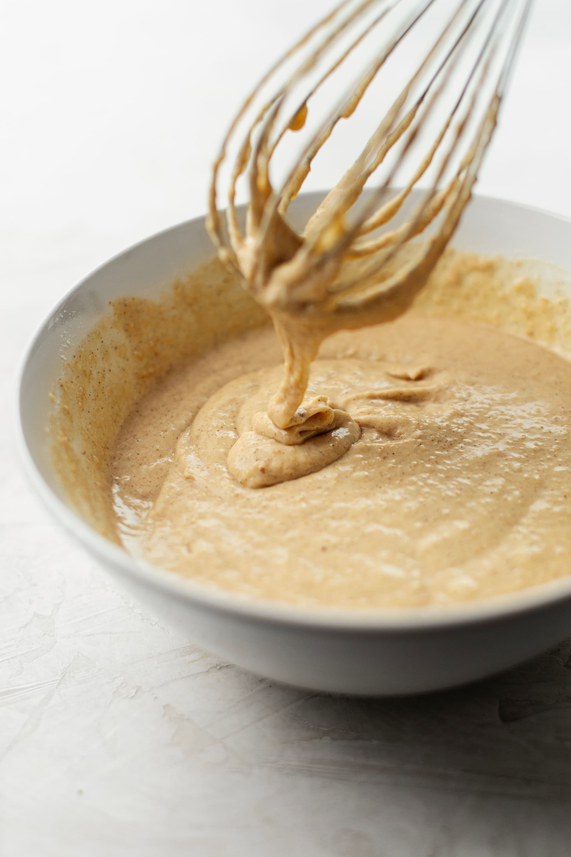 Almond cake batter dripping from a whisk, smooth in texture and warm golden-brown in colour.